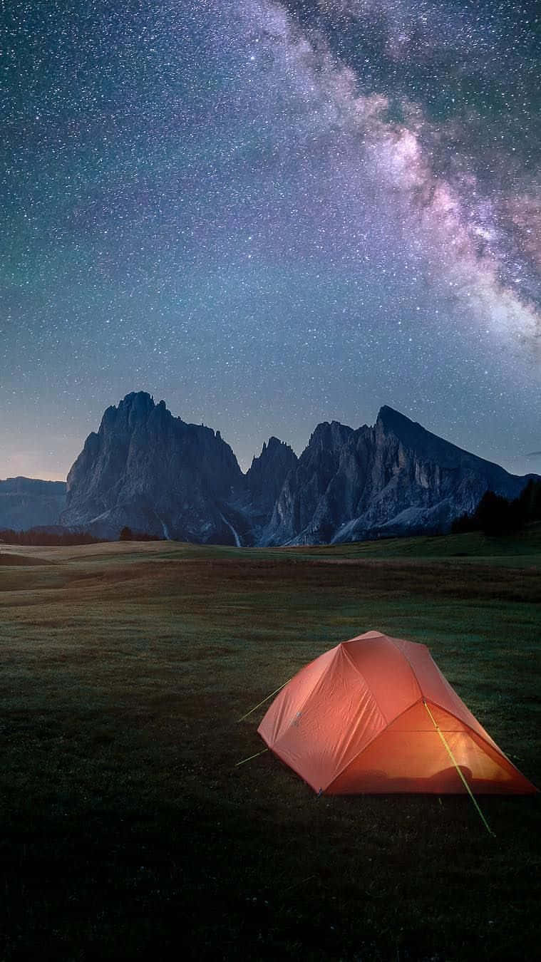 山景 桌布 壁紙 — 一個發光的橙色帳篷獨自矗立在草地上的草地上，下方是令人嘆為觀止的星空，銀河在崎嶇的山脈上空拱起。