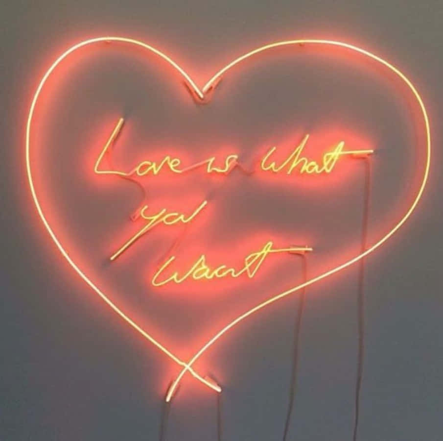 Orange Heart 桌布 壁紙 — 一個發光的橙色心形霓虹燈，上面有草書文字 'Love is what you want'，背景是柔和的灰色牆壁。