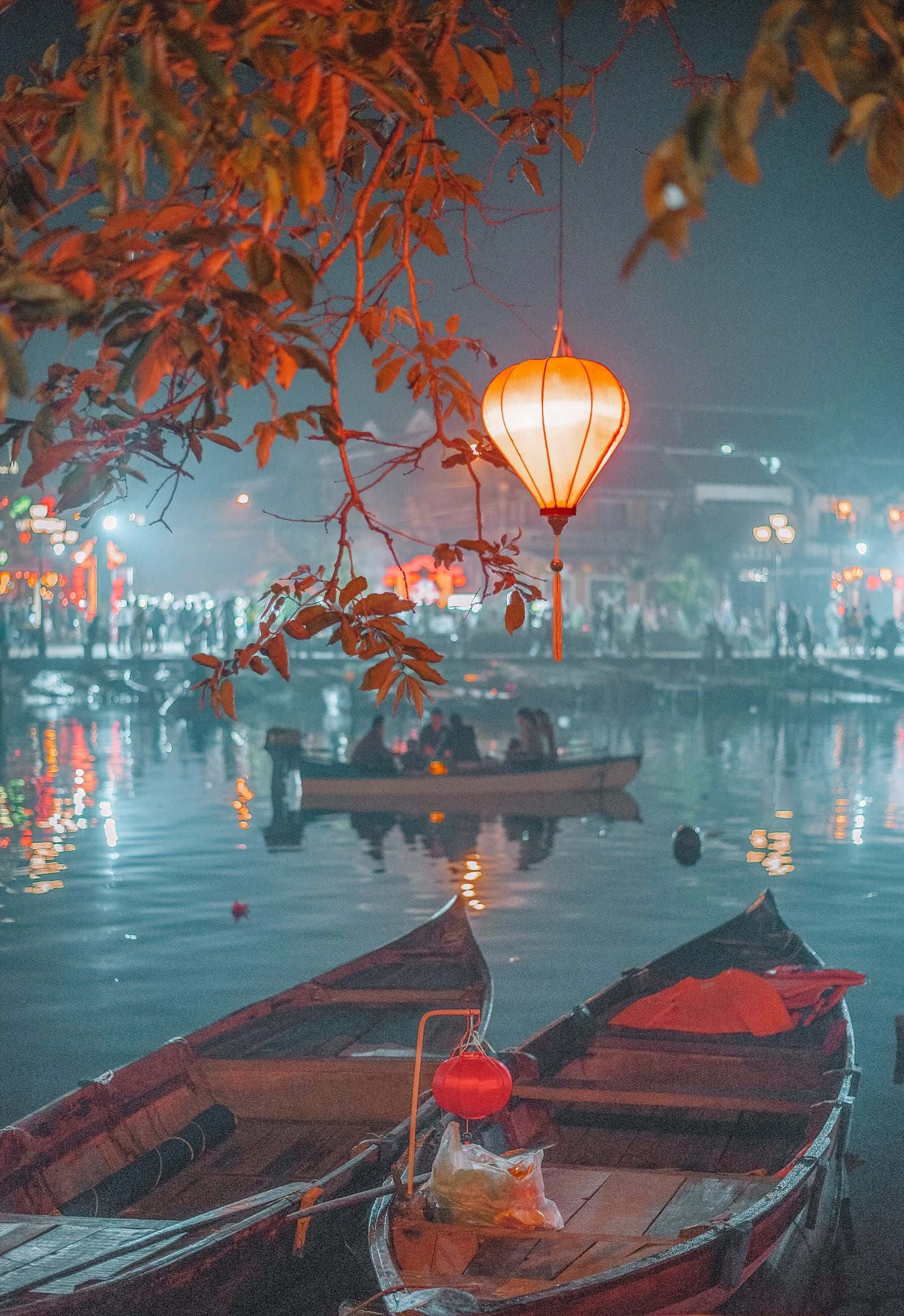 越南 桌布 壁紙 — 一個發光的橙色燈籠掛在秋天的樹葉上，下方是霧氣瀰漫的夜湖上的木船，遠處的燈光倒映在水面上。