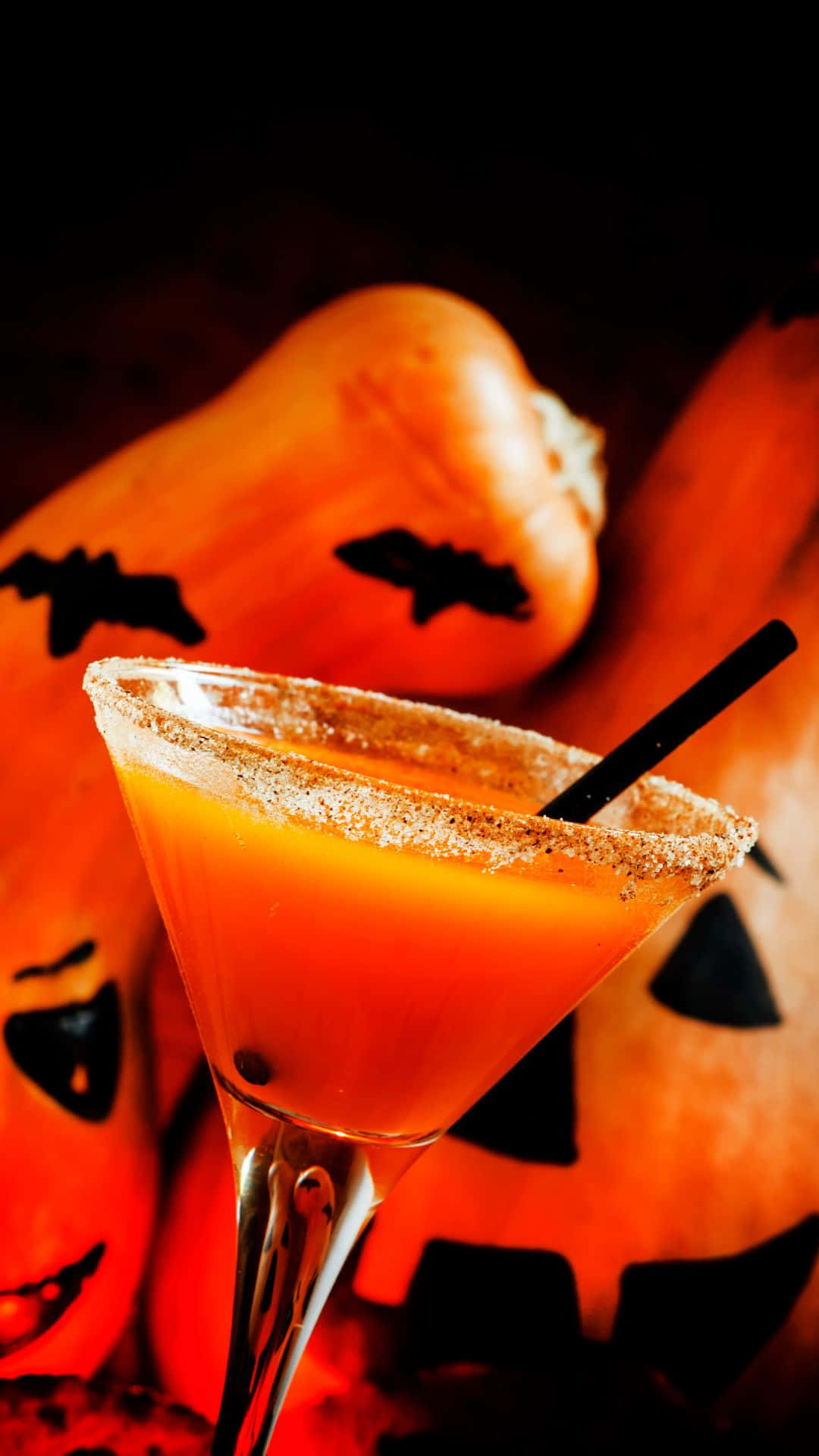 Spooky Cocktails 桌布 壁紙 — 一個發光的橙色萬聖節雞尾酒，放在糖邊馬丁尼杯中，前面是雕刻南瓜和蝙蝠剪影。