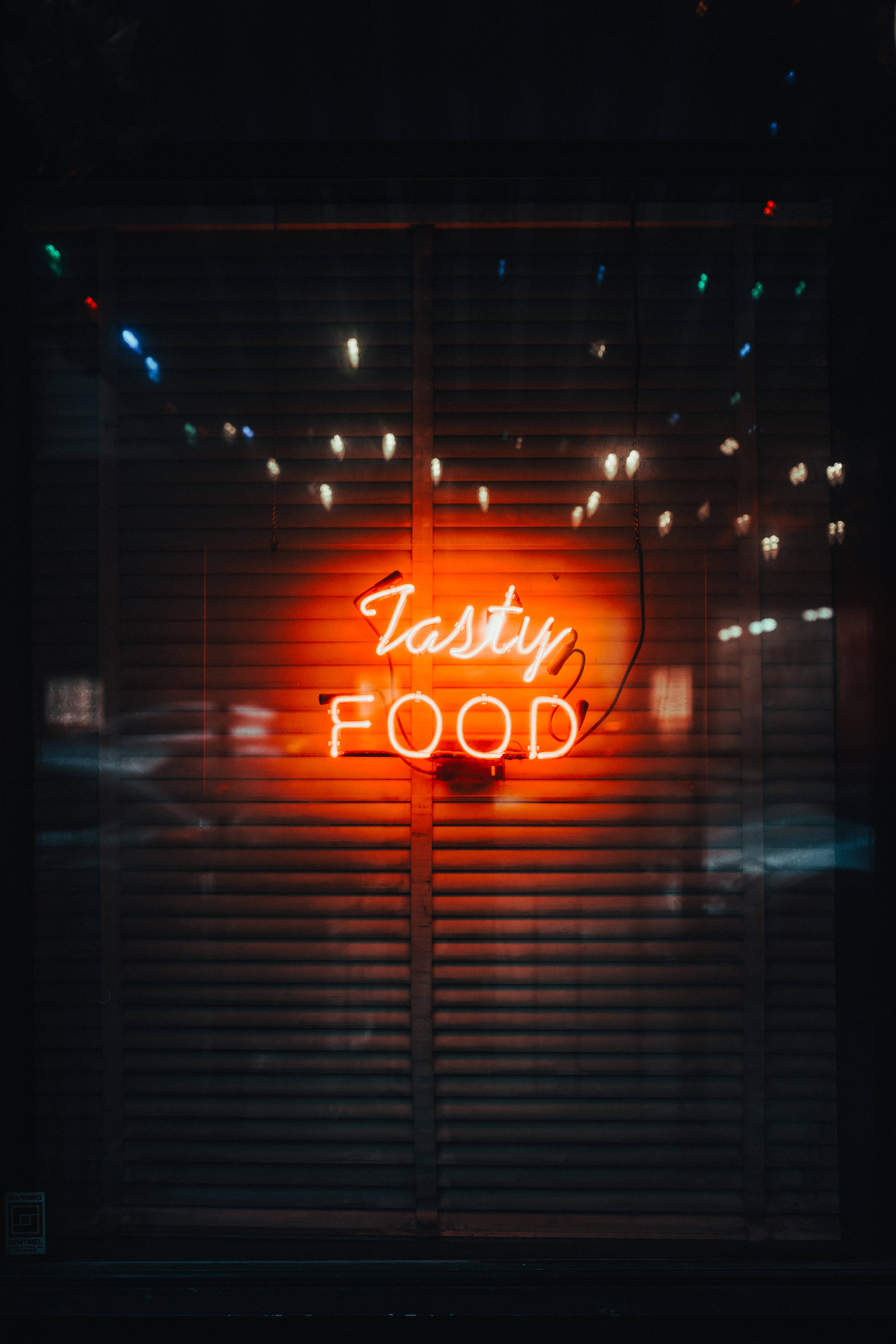 霓虹手機 桌布 壁紙 — 一個發光的橙色霓虹燈牌，上面寫著「Tasty FOOD」，穿過昏暗城市窗戶中的深色水平百葉窗照射出來。