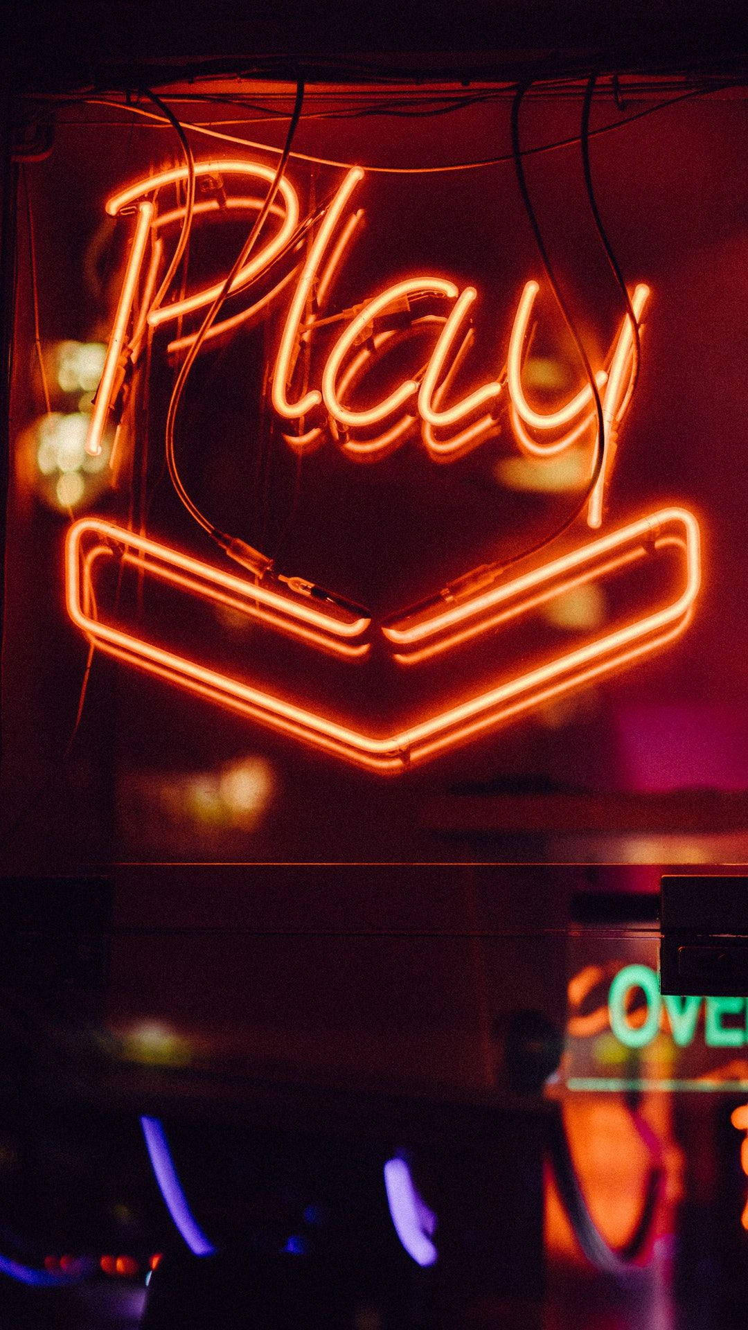 Neon Orange 桌布 壁紙 — 一個發光的橙色霓虹燈牌，拼出「Play」字樣並帶有向下箭頭，在昏暗的城市環境中投射出溫暖的光芒。