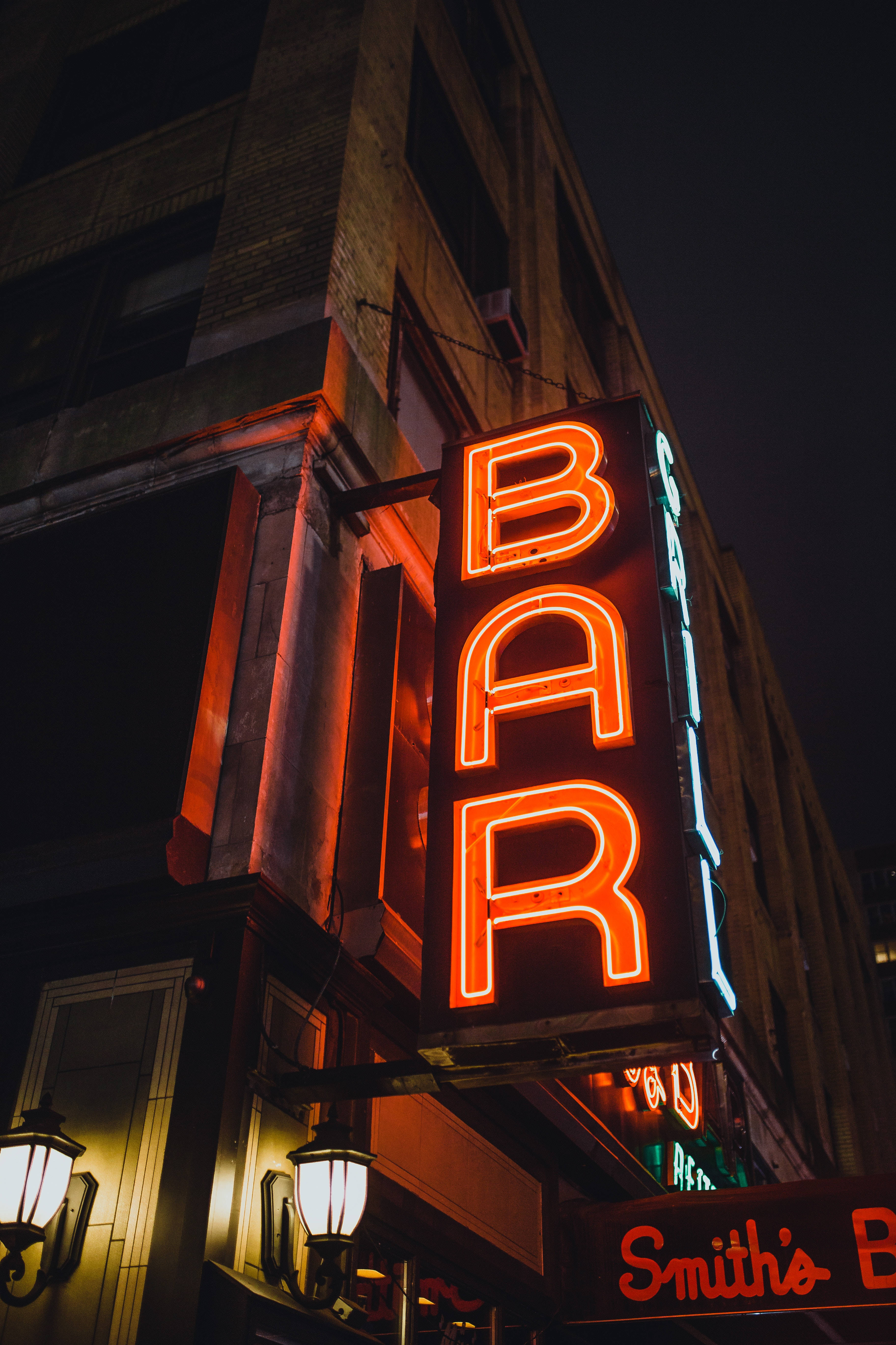 neon sign 桌布 壁紙 — 一個發光的橙色霓虹燈酒吧招牌在夜間照亮了磚砌建築的外牆，為周圍的建築物投下溫暖的光芒。