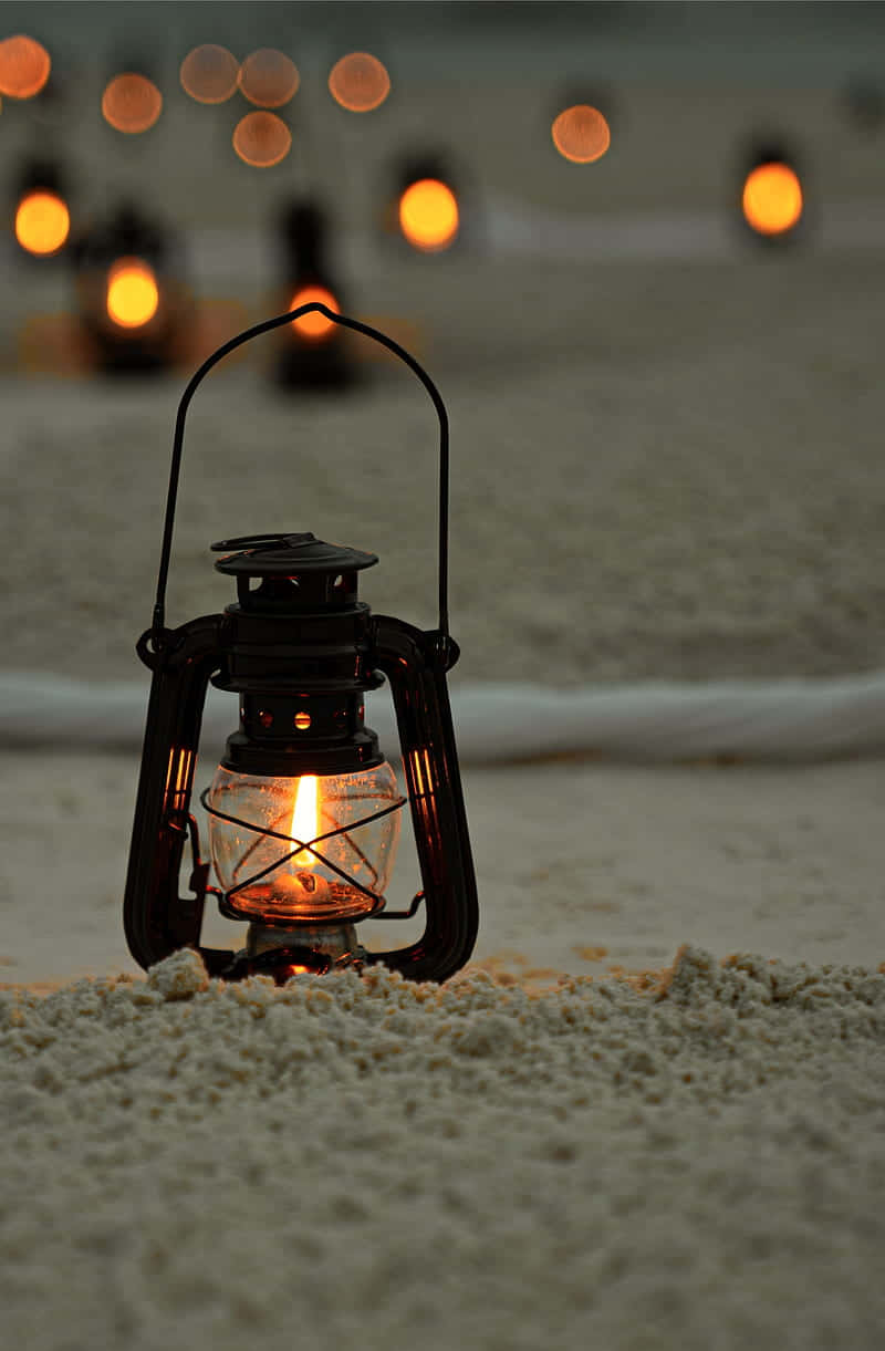 Lantern 桌布 壁紙 — 一個發光的油燈放在沙灘上，背景是黃昏時模糊的溫暖燈光。