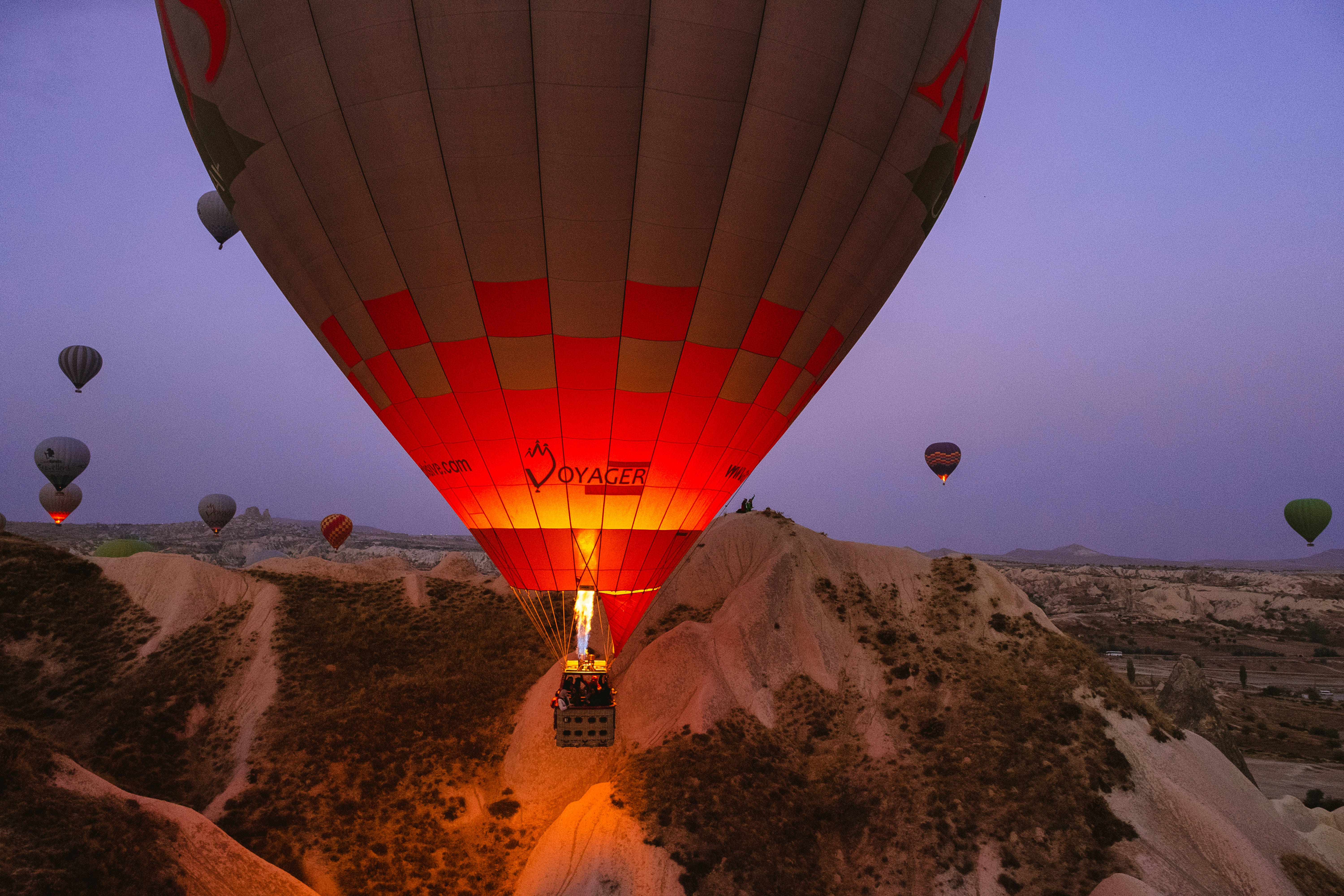 Cappadocia 桌布 壁紙 — 一個發光的熱氣球，帶有紅色和米色面板，在黃昏時分升越崎嶇的岩石地貌，其他氣球在昏暗的天空中飄蕩。
