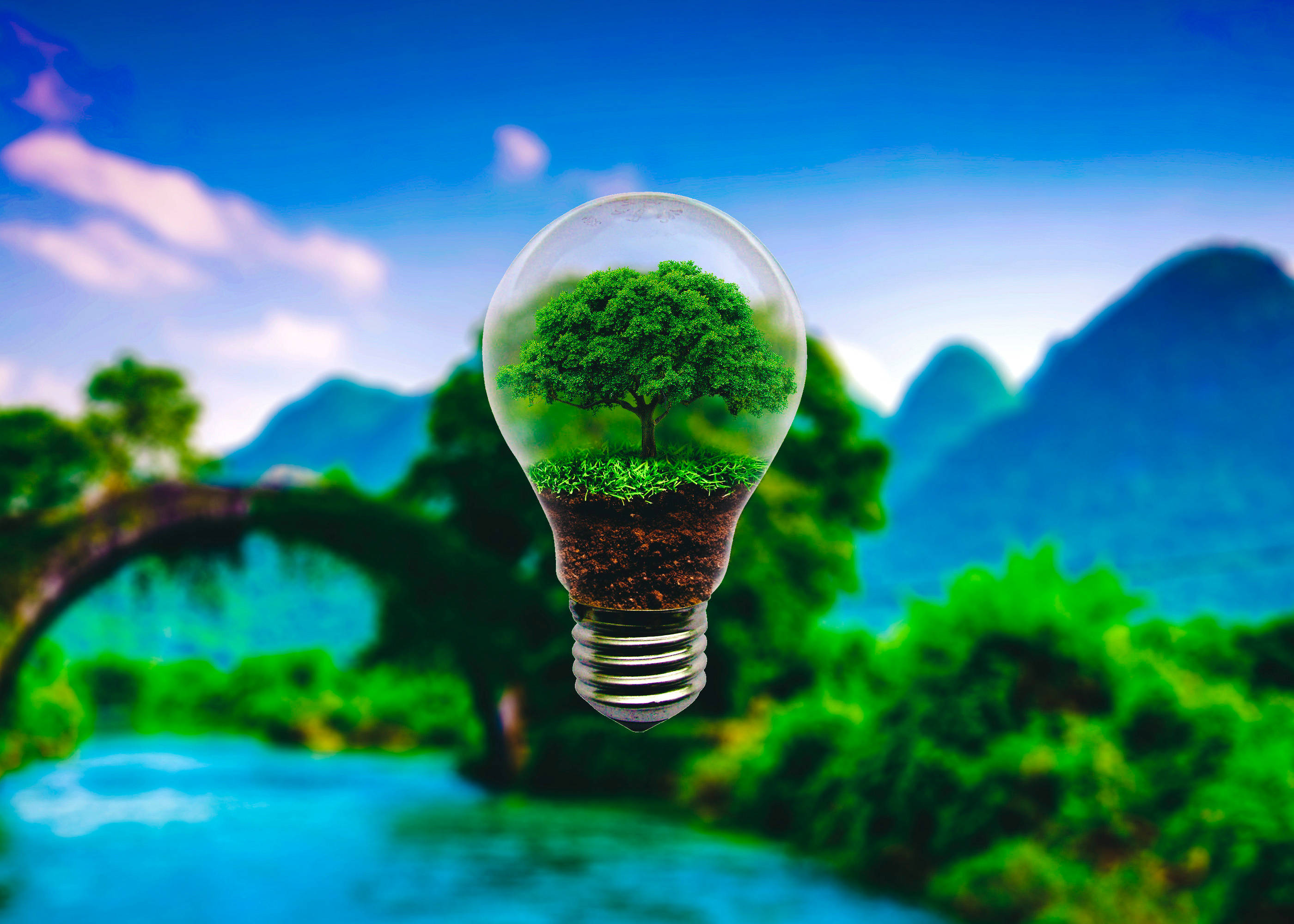Greenery 桌布 壁紙 — 一個發光的燈泡內含一棵茂盛的綠色樹木和泥土，背景是湛藍的天空和山巒景觀。