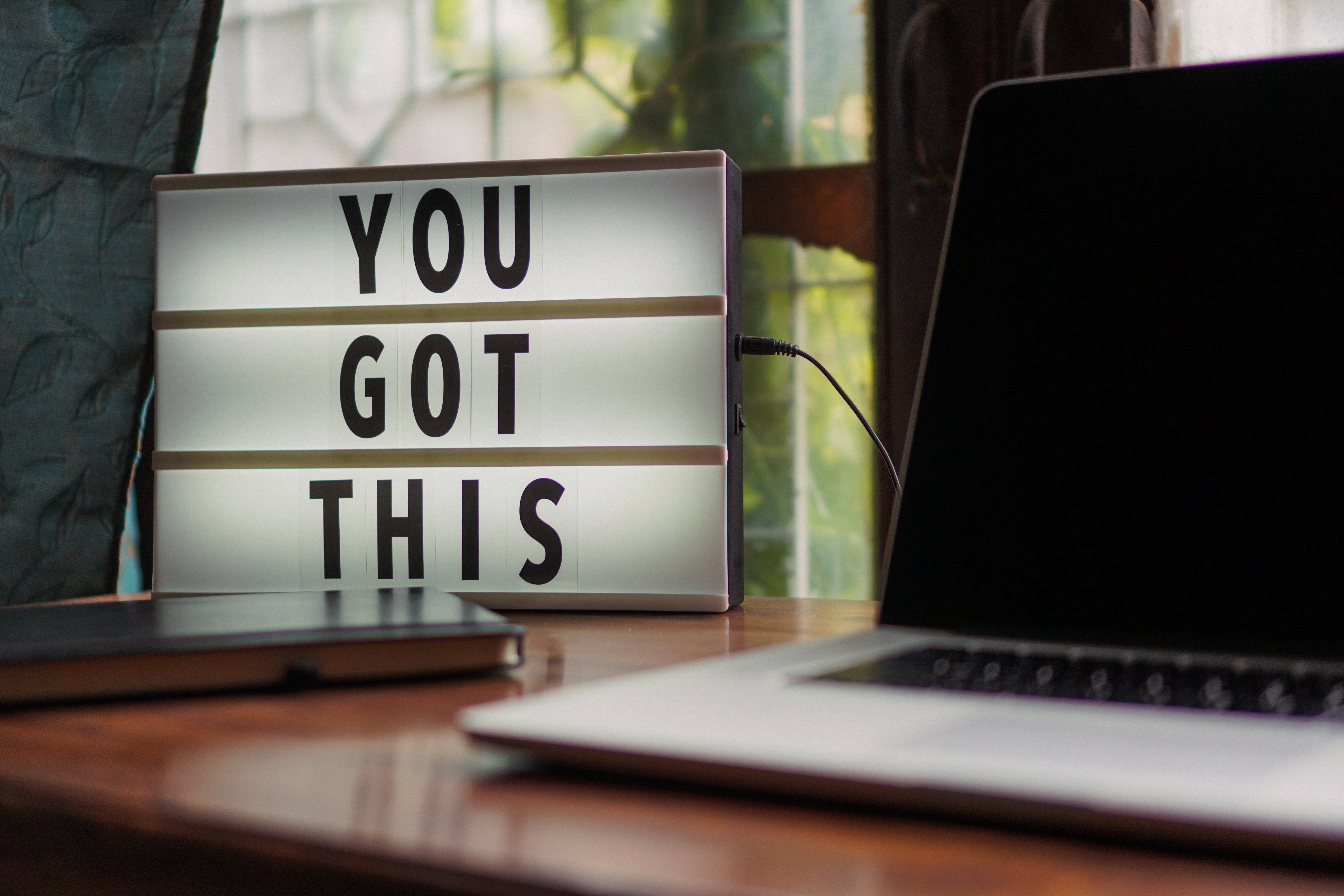 aesthetic tumblr laptop 桌布 壁紙 — 一個發光的燈箱，上面寫著「YOU GOT THIS」，放在木桌上，旁邊有一部筆記型電腦，靠近有綠色植物的窗戶。