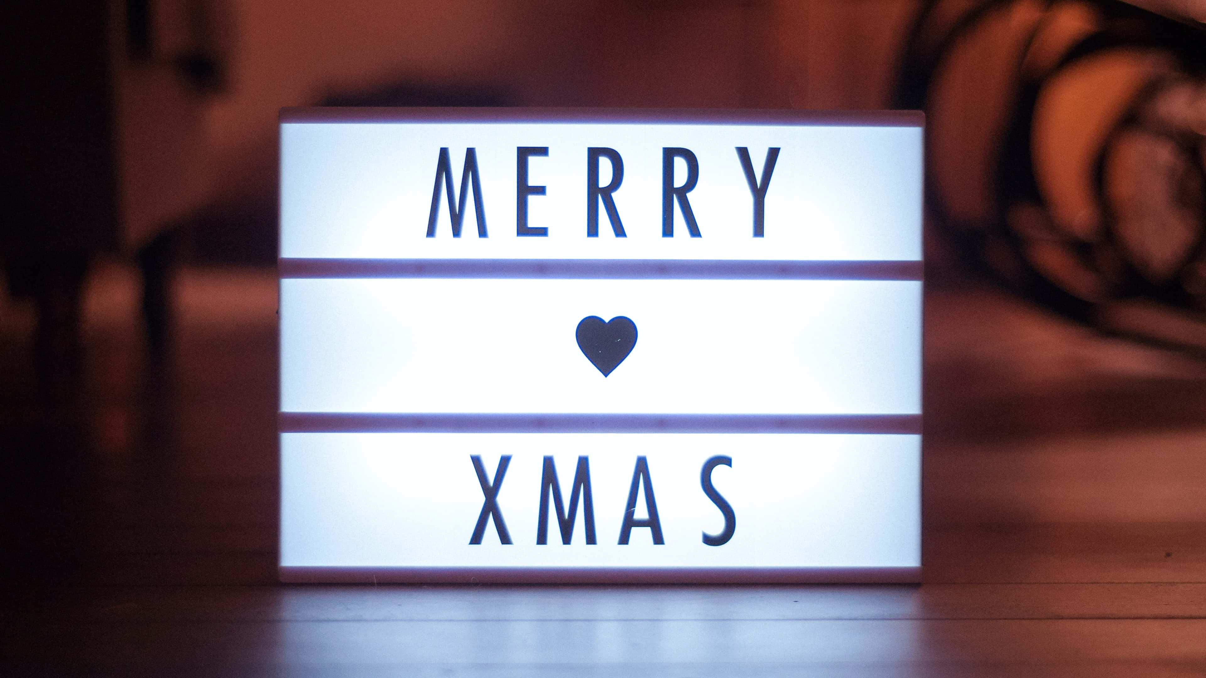節日 桌布 壁紙 — 一個發光的燈箱在溫暖模糊的木質背景上顯示「MERRY XMAS」和一個心形符號。