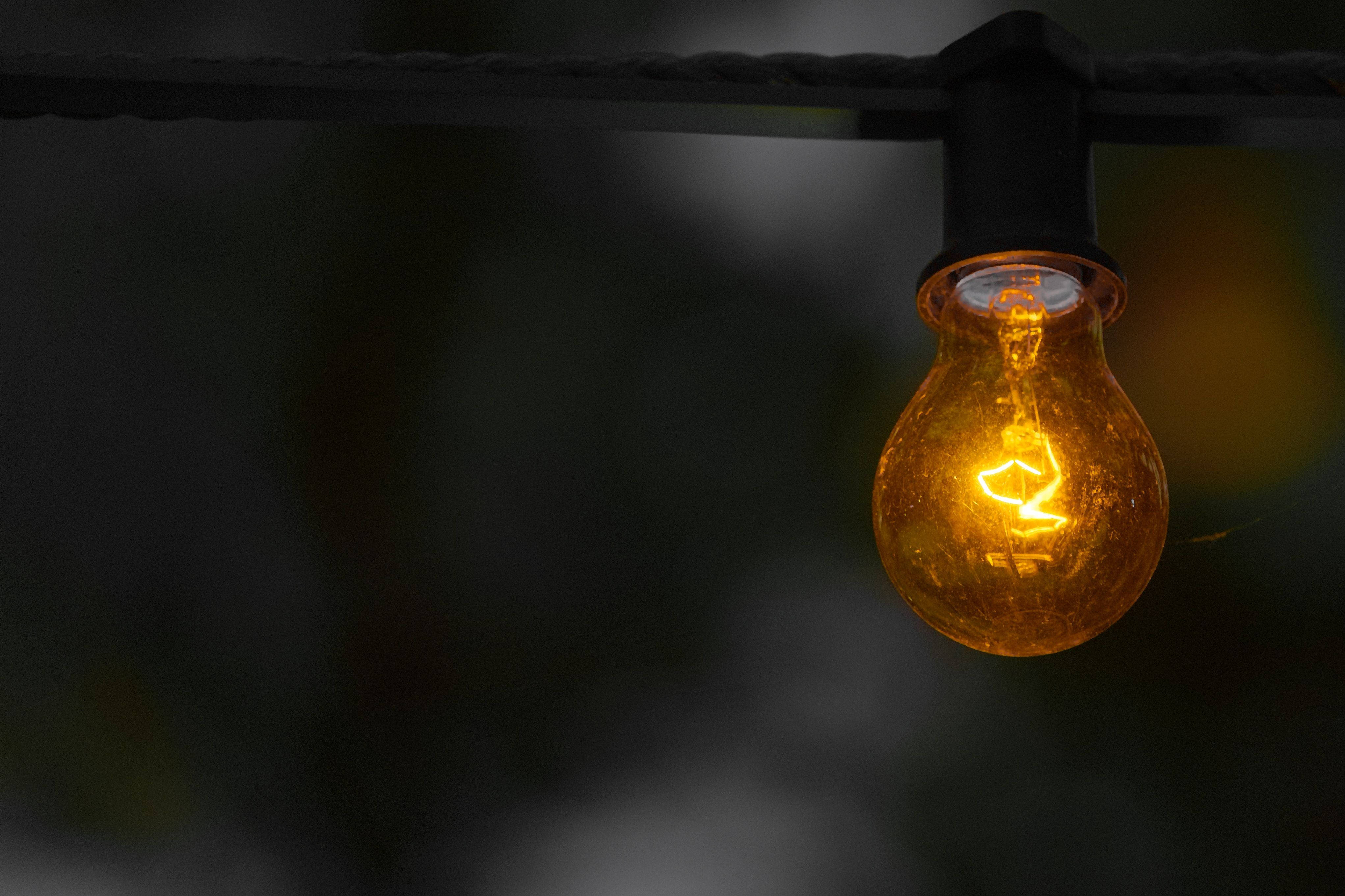 電力 桌布 壁紙 — 一個發光的琥珀色燈絲燈泡掛在黑色電線上，背景是黑暗模糊的。