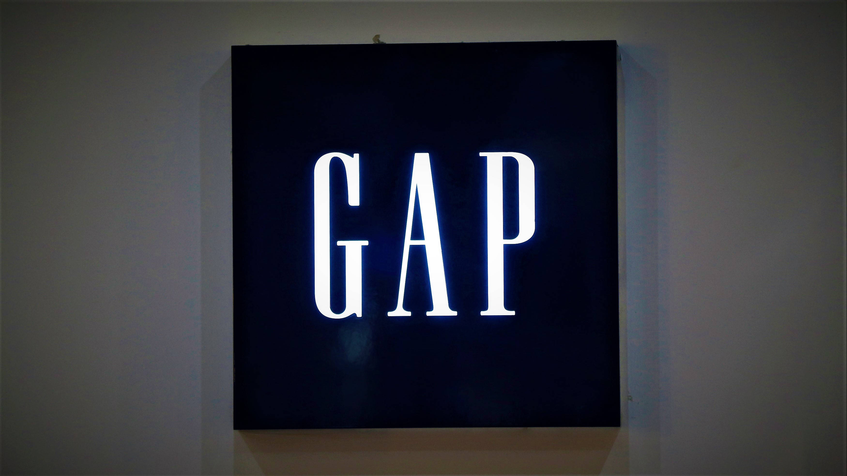 Gap 桌布 壁紙 — 一個發光的白色GAP標誌招牌，安裝在昏暗室內空間的素牆上。