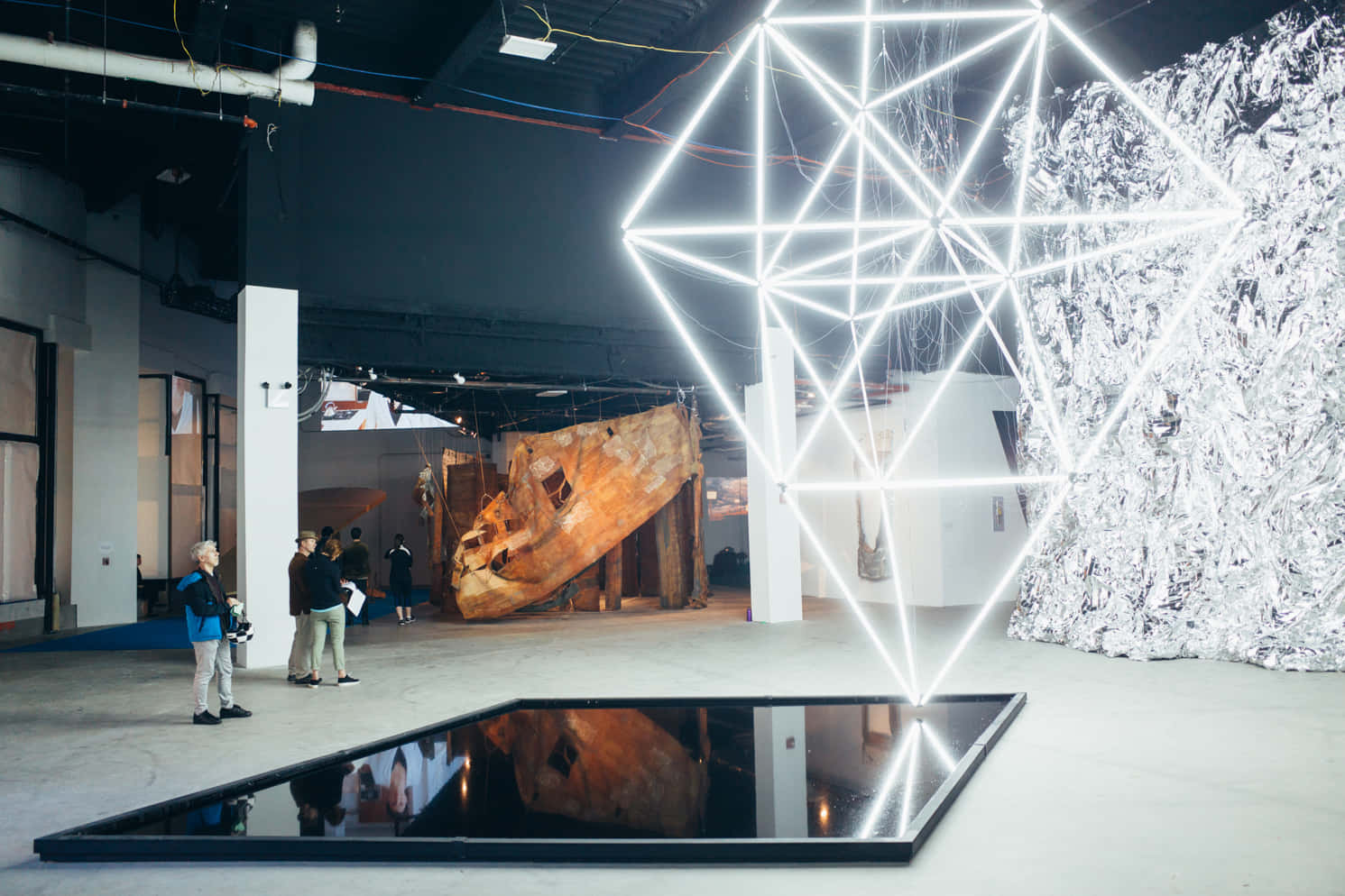 Installation Art 桌布 壁紙 — 一個發光的白色幾何光雕塑主導了現代畫廊空間，參觀者正在觀賞包括生鏽船體和反射地板的藝術裝置。
