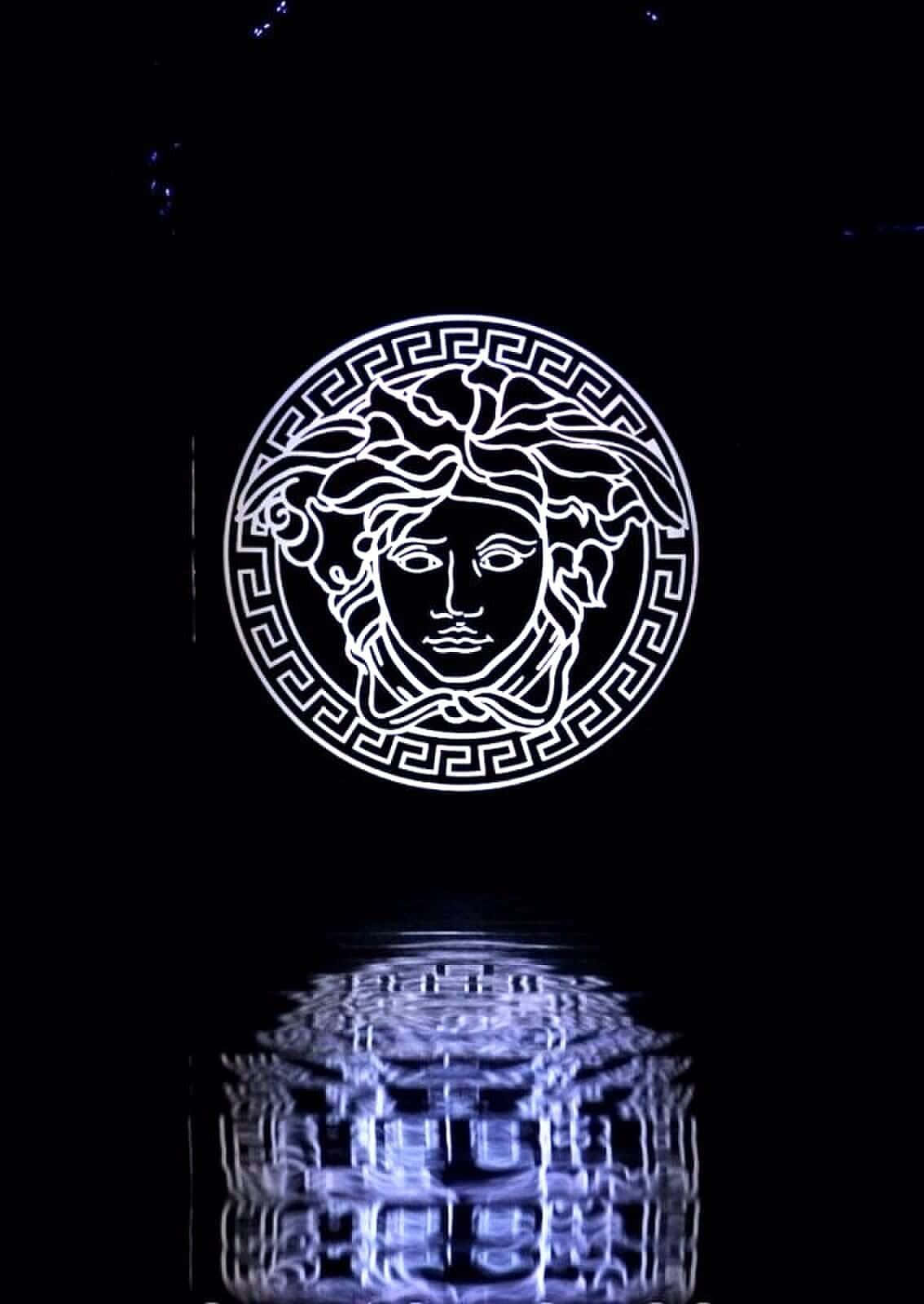Luxury 桌布 壁紙 — 一個發光的白色美杜莎頭標誌，帶有希臘鑰匙邊框，漂浮在黑暗水面上扭曲的藍色倒影之上。