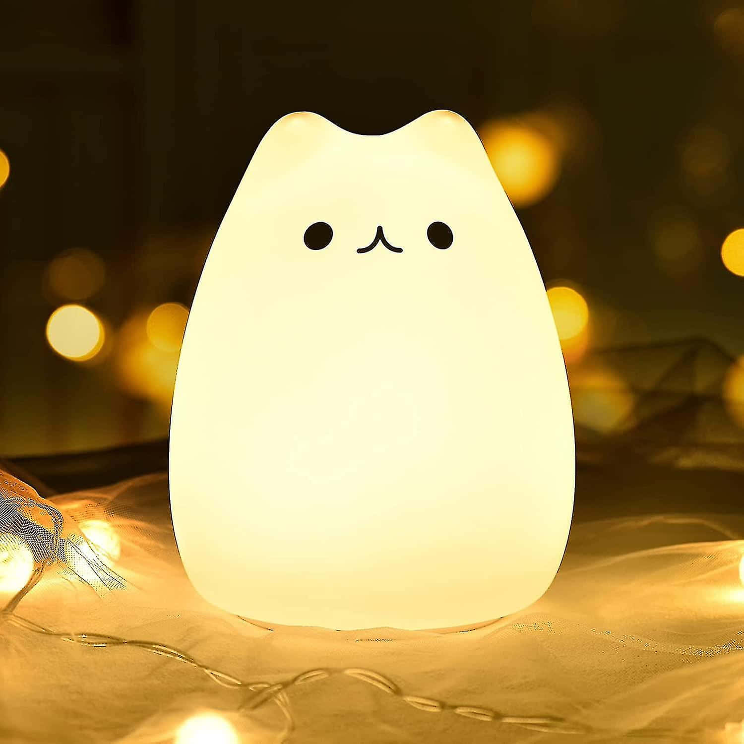 cute kitty 桌布 壁紙 — 一個發光的白色貓形燈，帶有簡單的黑色面部特徵，放置在柔軟的表面上，周圍環繞著溫暖的散景燈光。