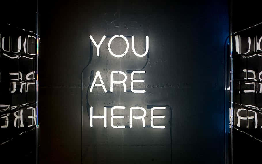 Tumblr White 桌布 壁紙 — 一個發光的白色霓虹燈牌，上面寫著 YOU ARE HERE，在兩側有鏡面反射的深色紋理牆壁上投射出柔和的光線。
