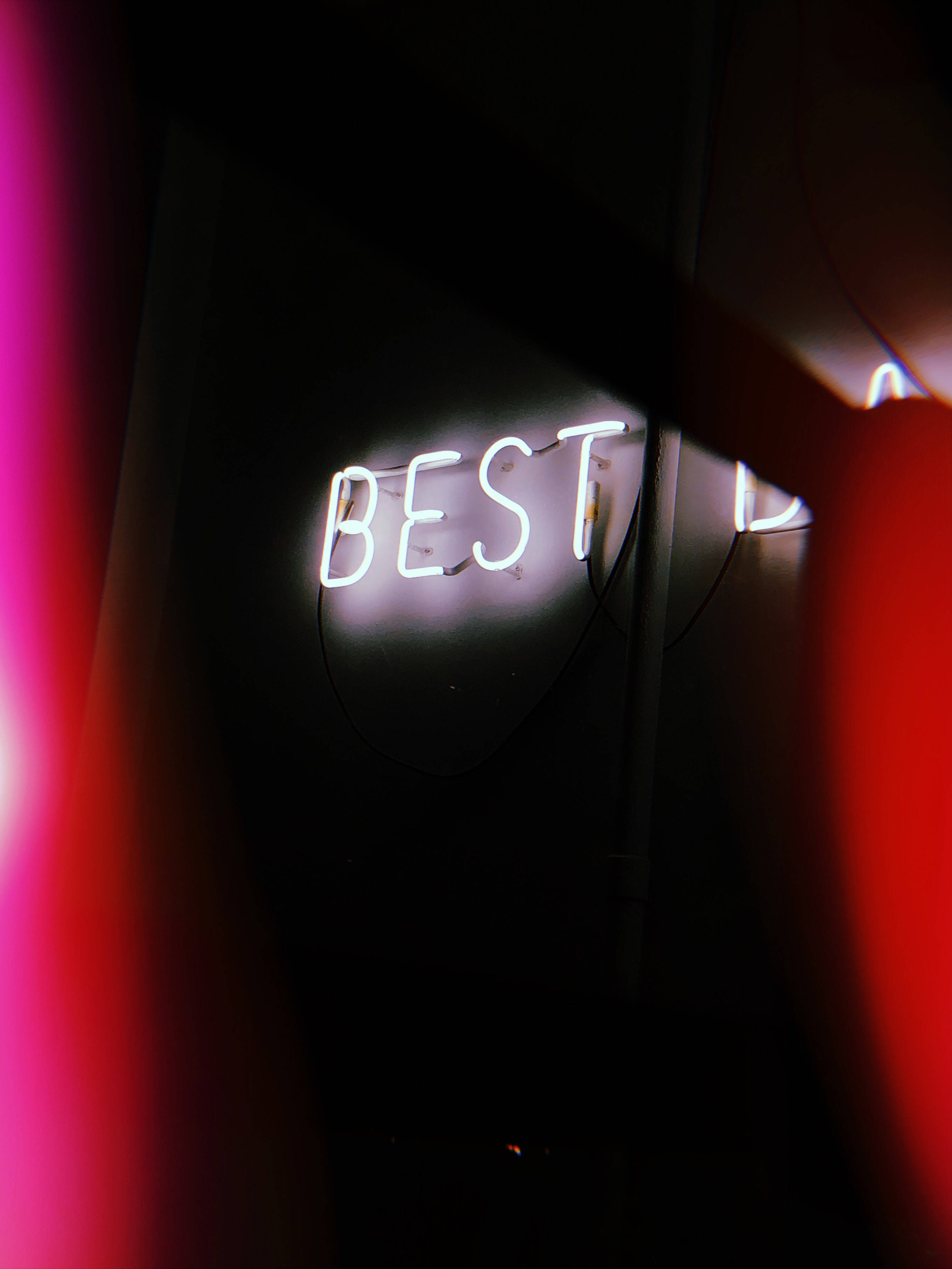 Best 桌布 壁紙 — 一個發光的白色霓虹燈牌，拼出「BEST」，照亮了一面被模糊的紅色和粉紅色光條紋框住的暗牆。