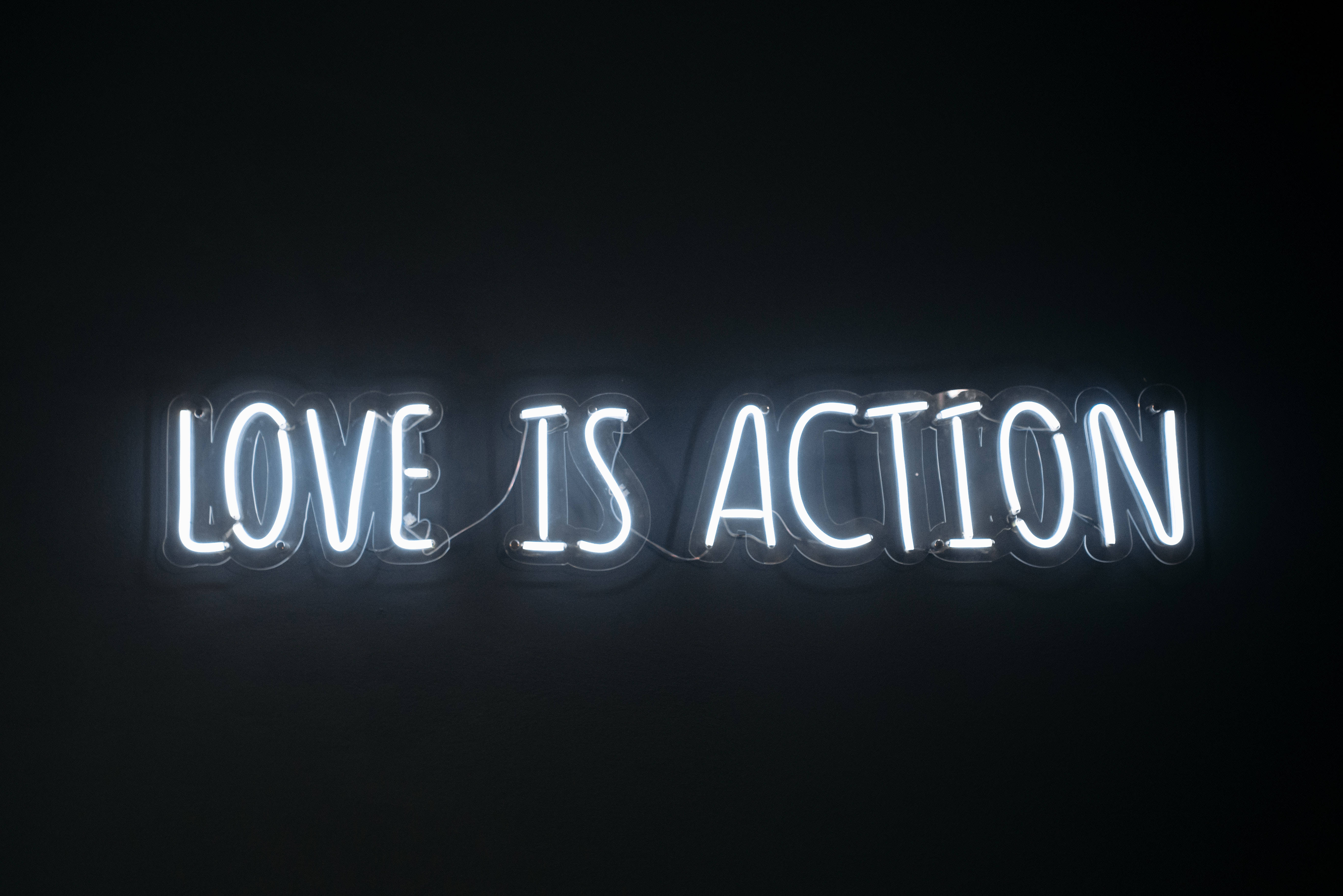 Word 桌布 壁紙 — 一個發光的白色霓虹燈牌在純黑背景上顯示「LOVE IS ACTION」。