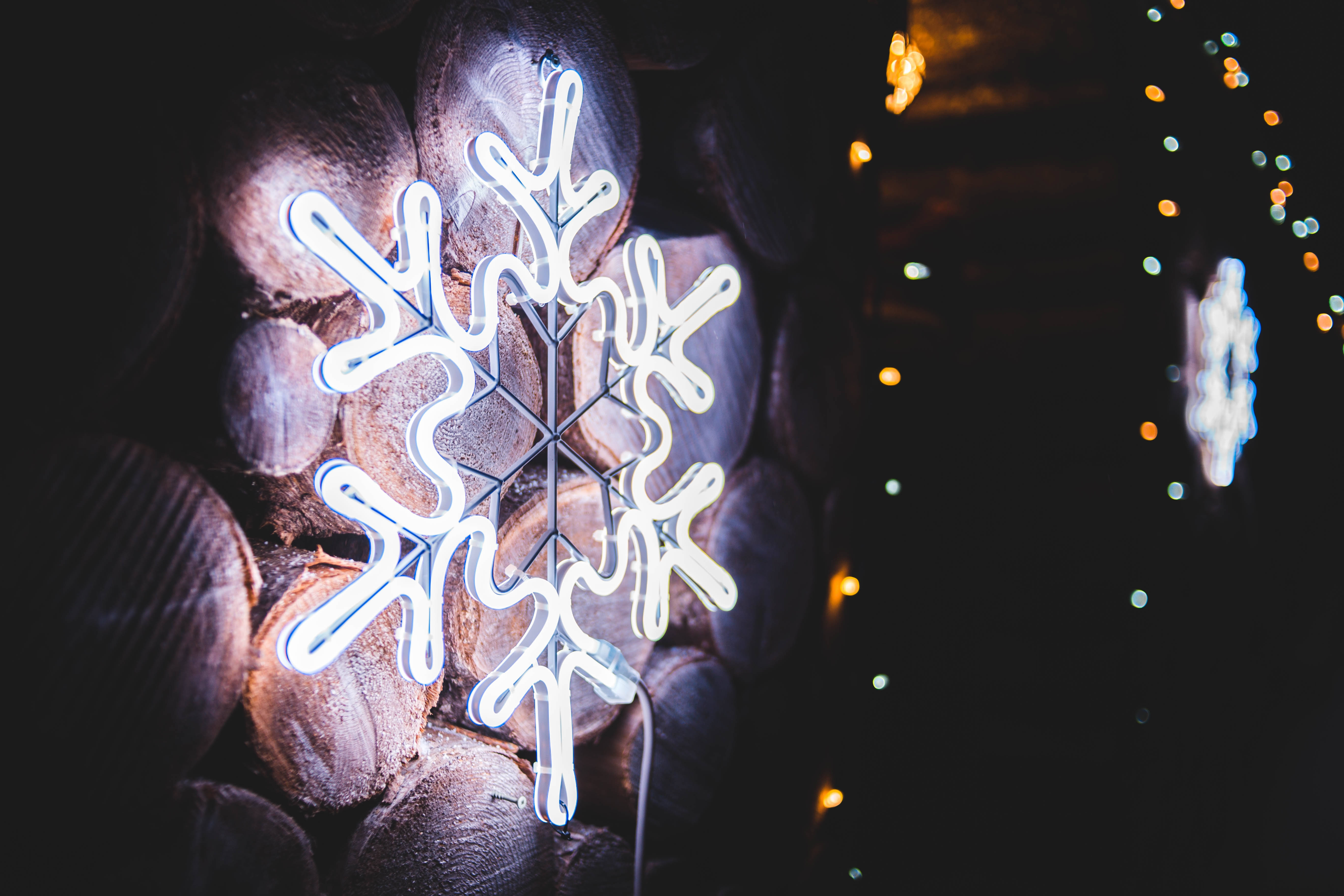 雪花 桌布 壁紙 — 一個發光的白色霓虹雪花標誌靠在堆疊的木頭上，背景有模糊的節日燈光。