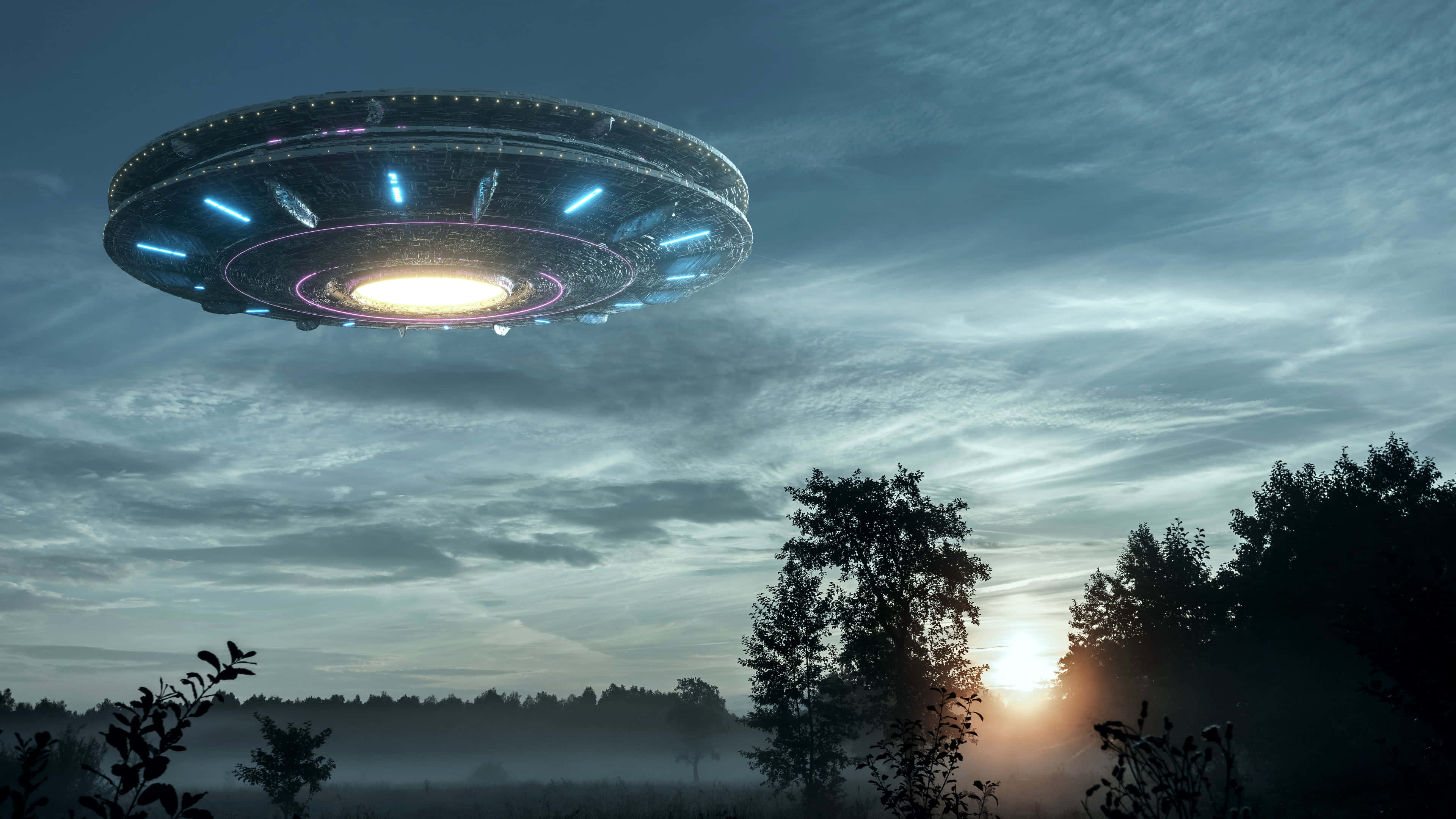 Real Ufo 桌布 壁紙 — 一個發光的碟形UFO在日落時分懸浮在霧氣籠罩的森林上空，向多雲的天空投射出藍色和白色的光芒。