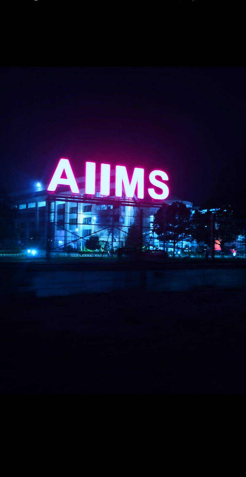 India 桌布 壁紙 — 一個發光的粉紅色AIIMS標誌在夜間照亮了一座現代建築，藍色燈光反射在潮濕的人行道上。