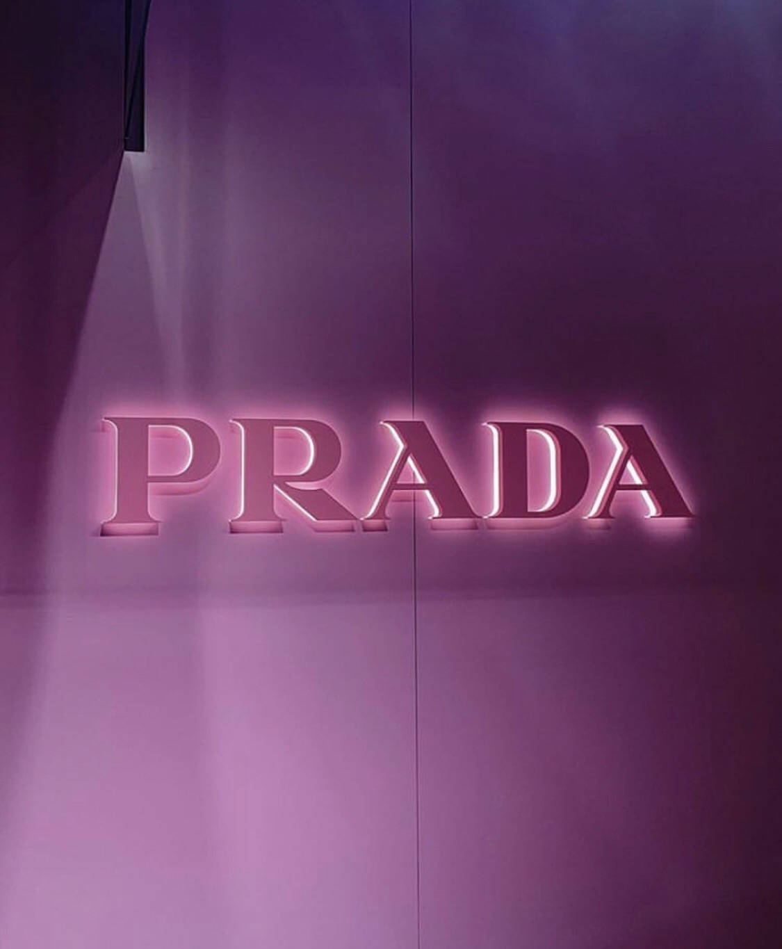 Prada 桌布 壁紙 — 一個發光的粉紅色Prada標誌招牌，安裝在光滑的紫色牆壁上，柔和燈光投射出輕柔陰影。