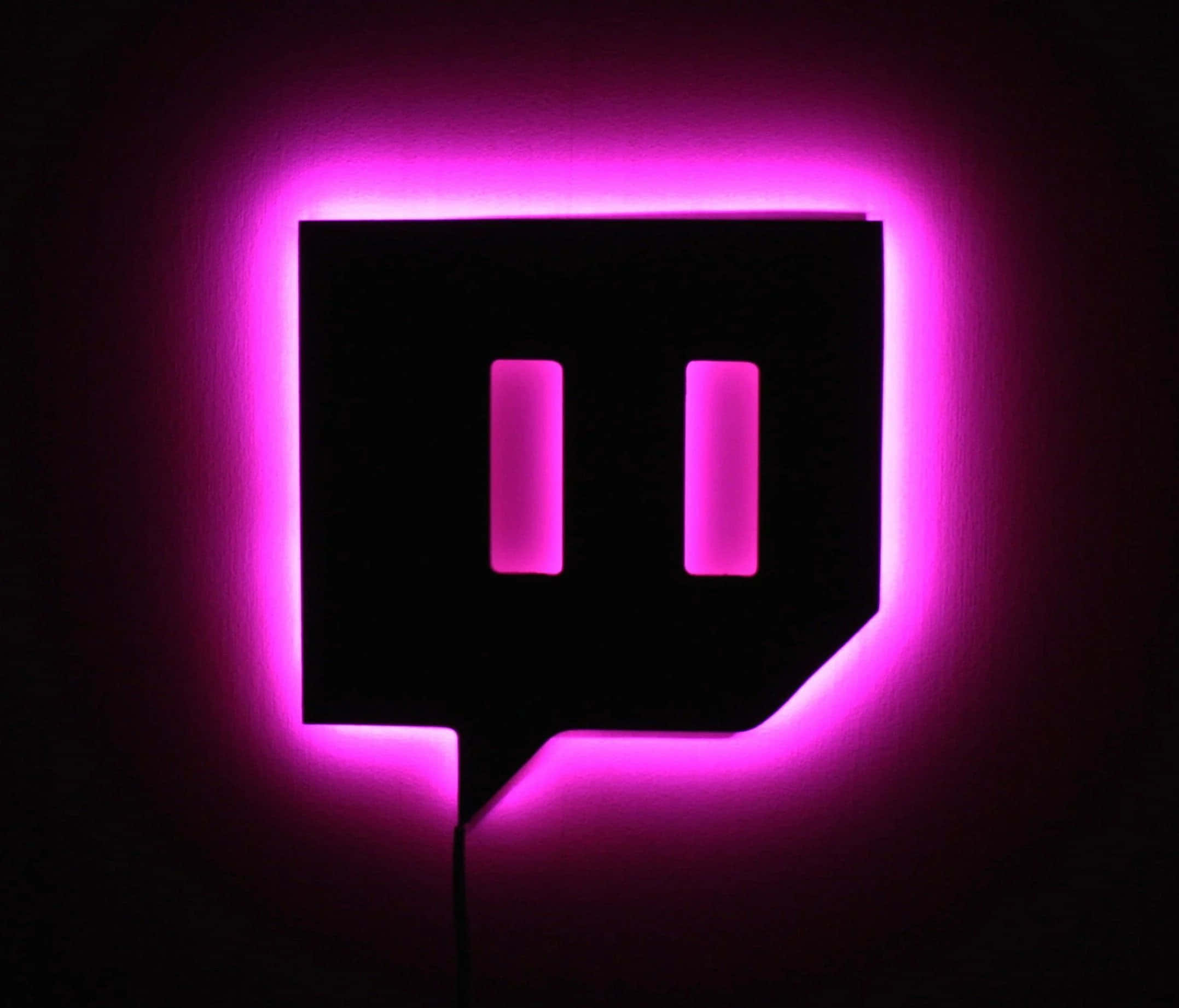 Twitch 桌布 壁紙 — 一個發光的粉紅色Twitch標誌招牌，帶有明亮的霓虹邊緣，掛在深色牆上。