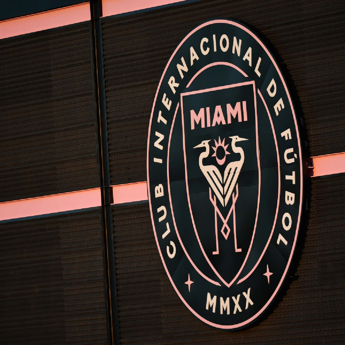 足球 桌布 壁紙 — 一個發光的粉紅色和黑色 Club Internacional de Fútbol Miami 標誌安裝在帶有水平燈條的深色紋理牆壁上。