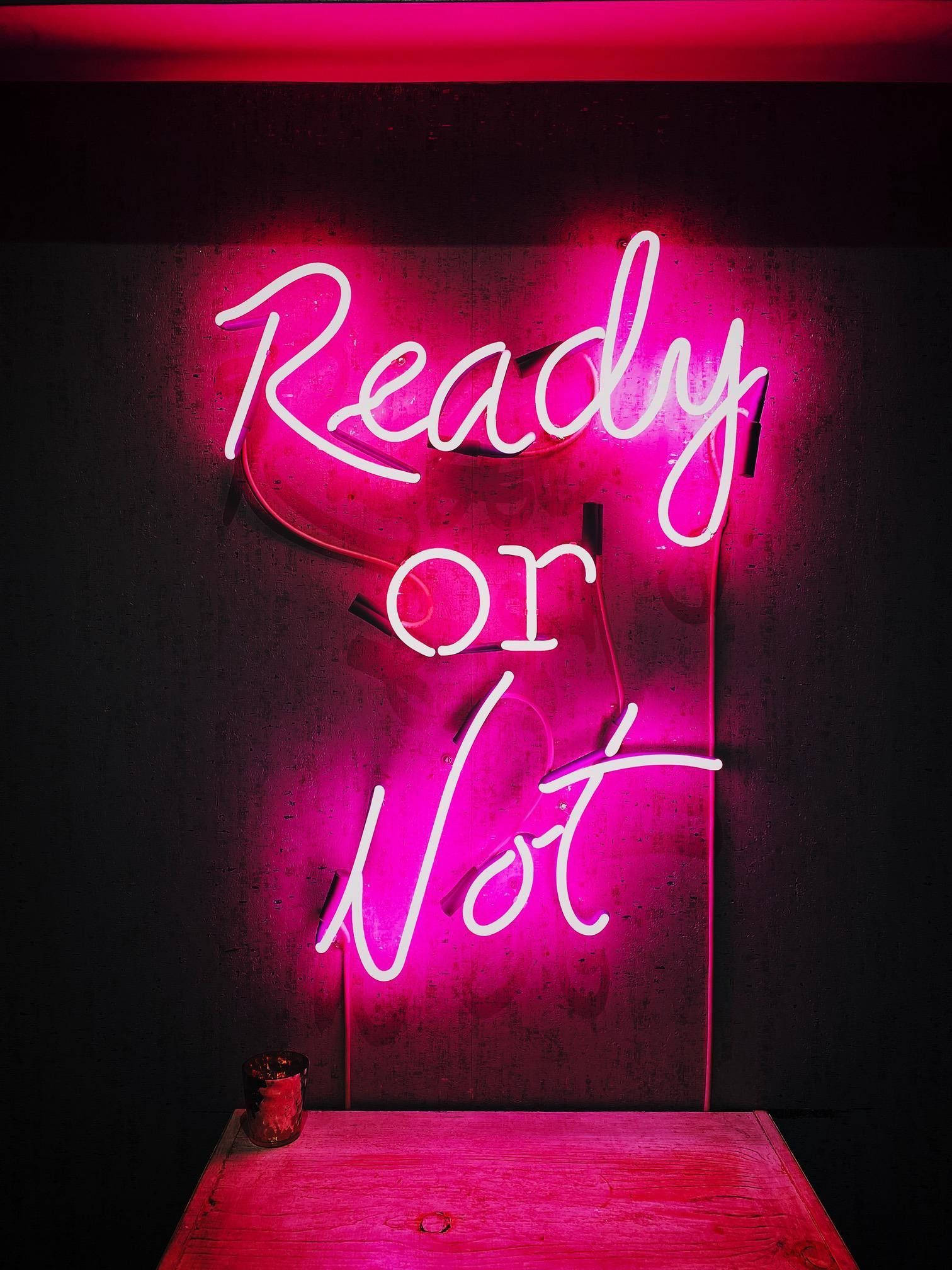 Neon Pink 桌布 壁紙 — 一個發光的粉紅色霓虹燈牌，上面寫著「Ready or Not」，在黑暗的牆壁和下面的木桌上投射出鮮豔的色彩。