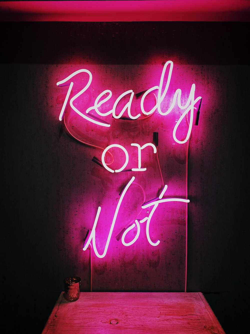 Hot Pink Aesthetic 桌布 壁紙 — 一個發光的粉紅色霓虹燈牌，上面寫著「Ready or Not」，在深色牆壁和下方的木桌上投射出鮮豔的色彩。