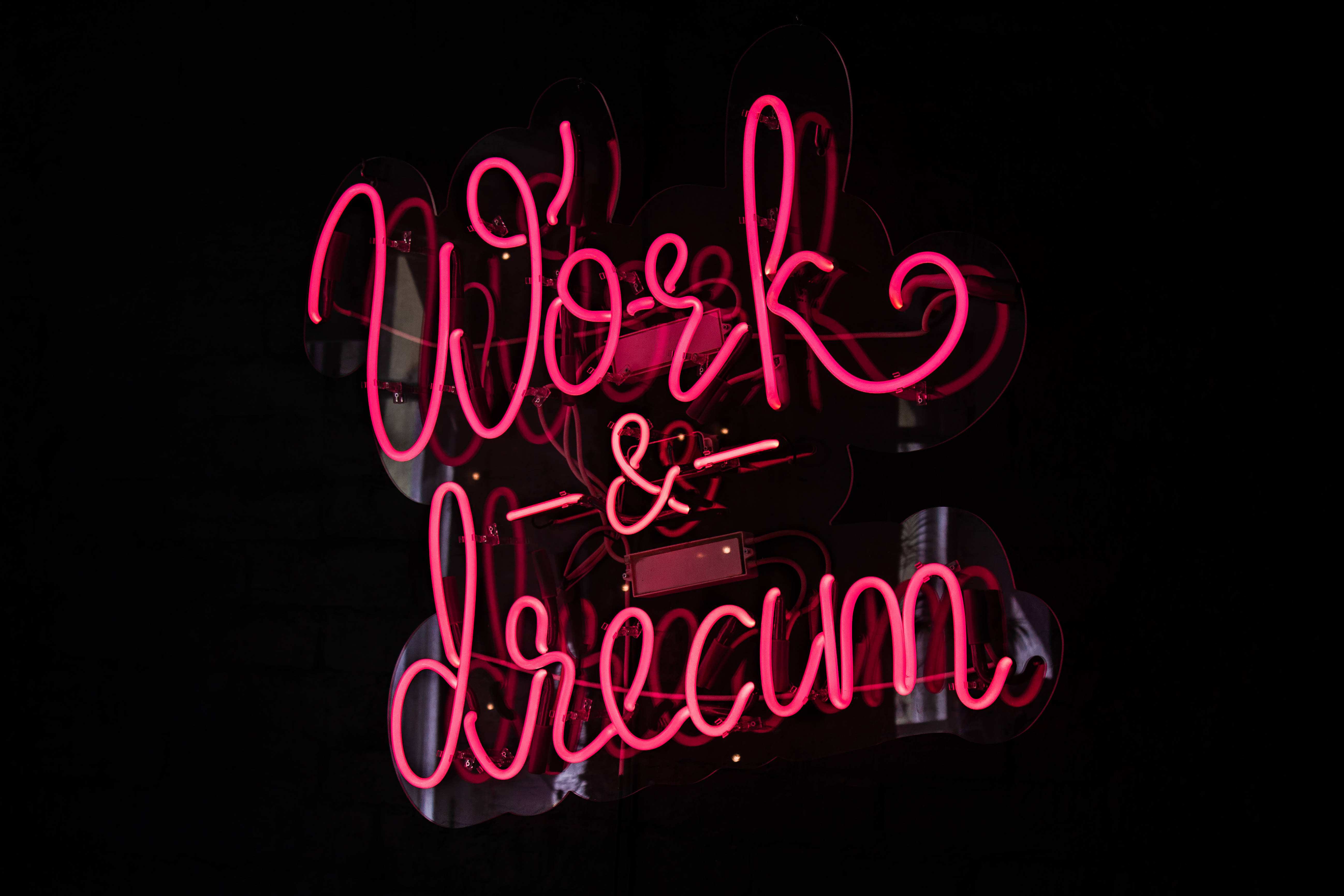 酷炫霓虹 桌布 壁紙 — 一個發光的粉紅色霓虹燈牌，上面寫著「Work & Dream」，背景為深色，並有可見的玻璃反射。