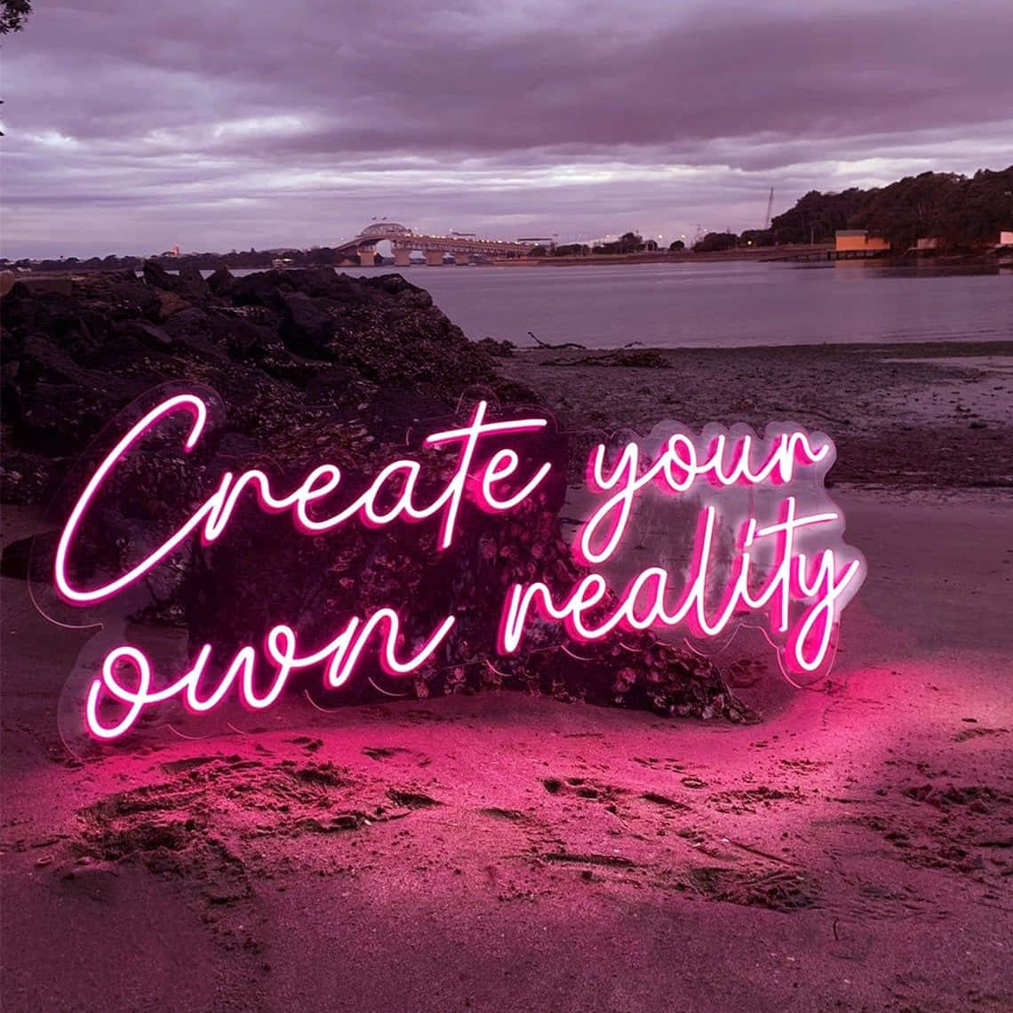 Neon Quotes 桌布 壁紙 — 一個發光的粉紅色霓虹燈牌，上面寫著「Create your own reality」，放在黃昏時分的岩石海灘上，遠處有一座橋，天空是憂鬱的紫色。