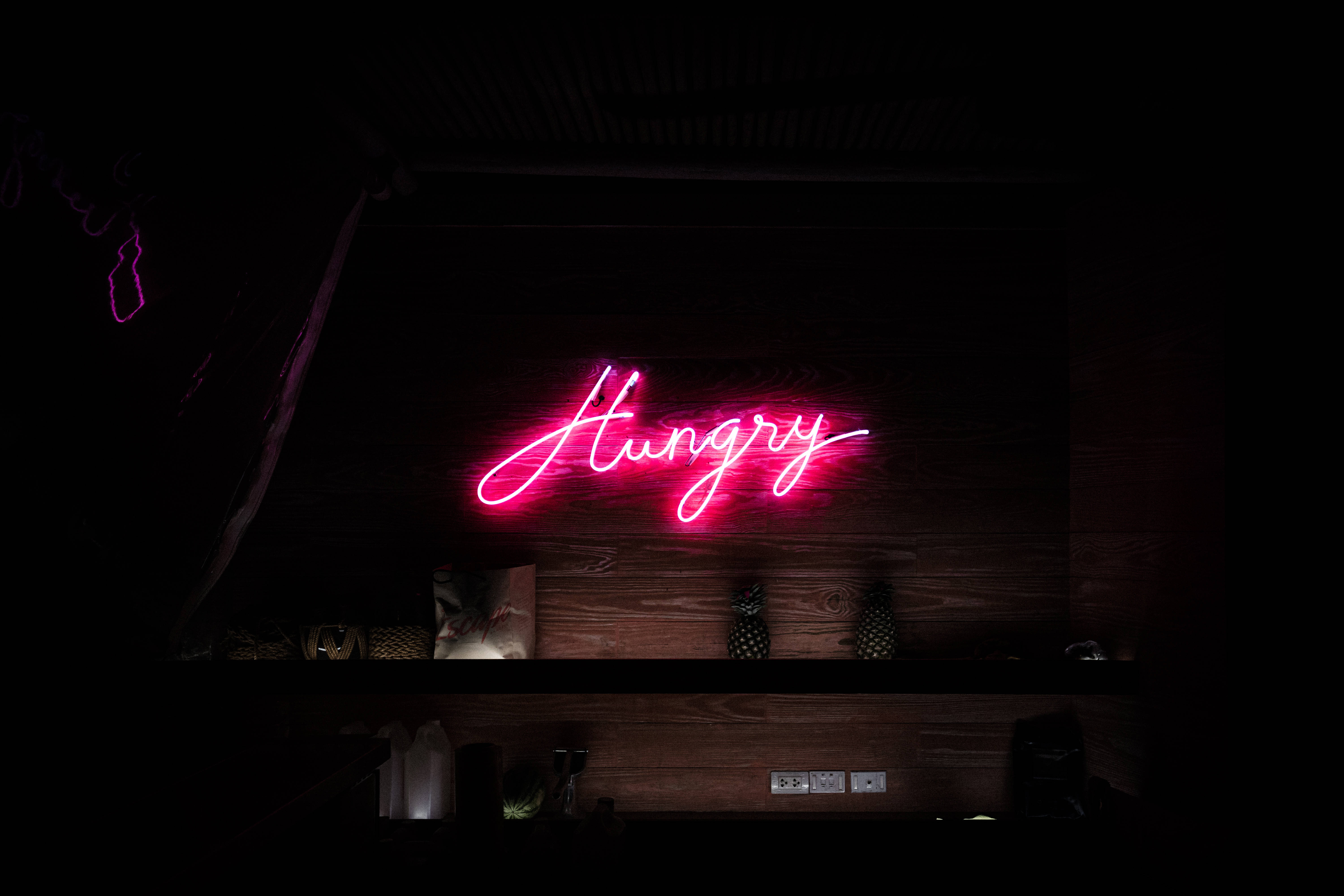 螢光黃 桌布 壁紙 — 一個發光的粉紅色霓虹燈牌，上面寫著「Hungry」，照亮了昏暗酒吧架子上方的深色木牆。