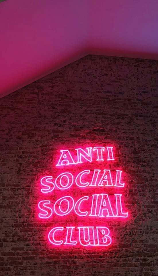 Anti Social Club iPhone 桌布 壁紙 — 一個發光的粉紅色霓虹燈牌，上面寫著「ANTI SOCIAL SOCIAL CLUB」，照亮了昏暗房間中帶有紋理的磚牆。
