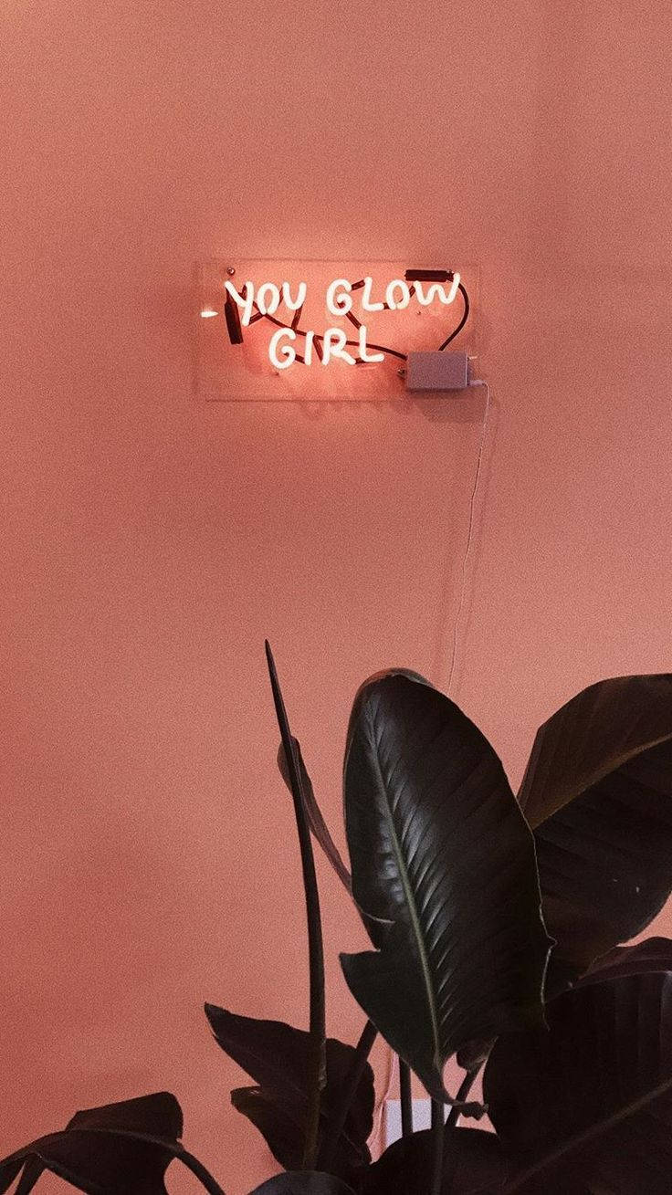 iPhone 桌布 壁紙 — 一個發光的粉紅色霓虹燈牌，上面寫著「YOU GLOW GIRL」，安裝在相配的粉紅色牆上，下方有深色熱帶葉子。