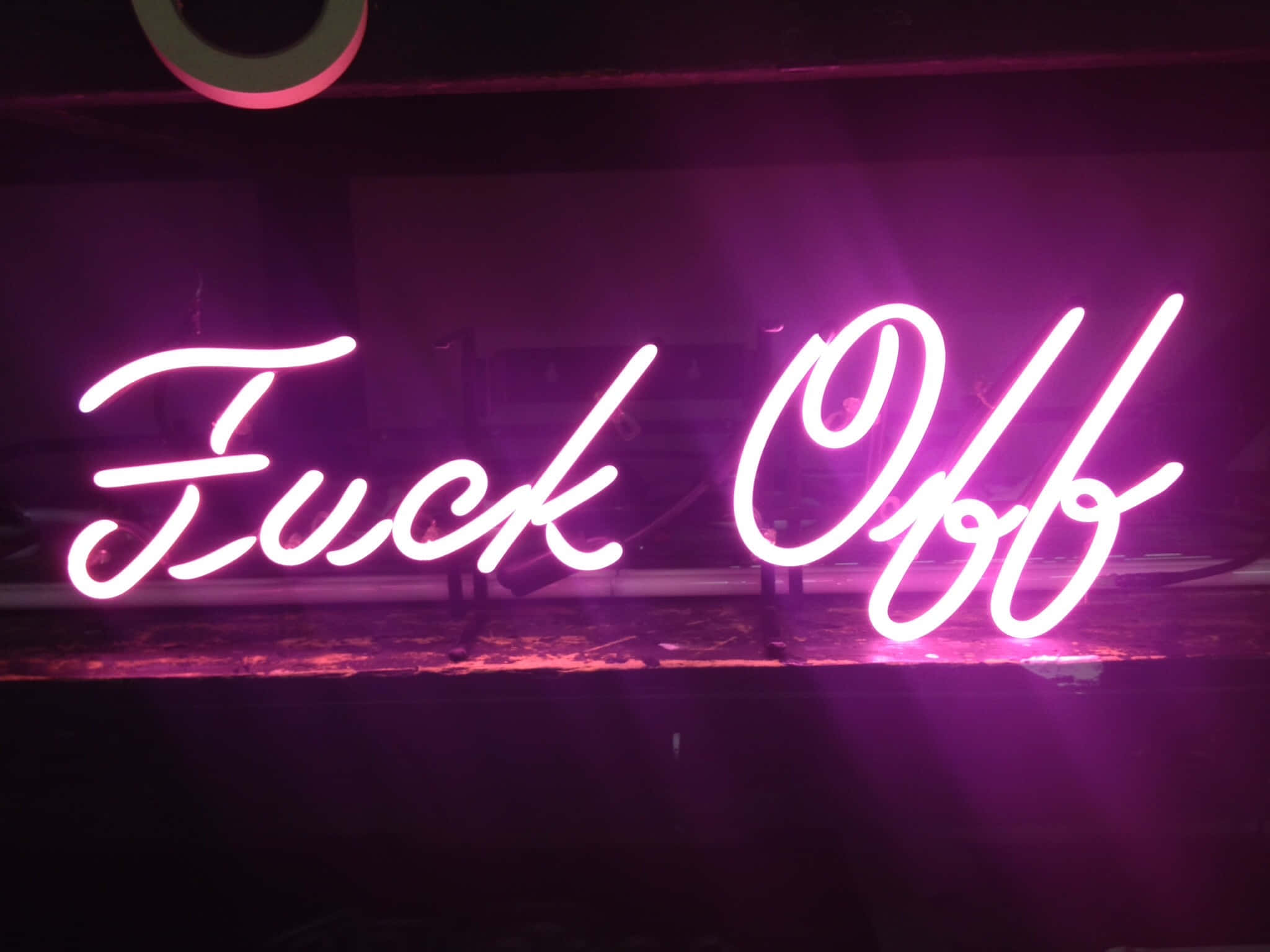 neon sign 桌布 壁紙 — 一個發光的粉紅色霓虹燈牌，上面有草書的「Fuck Off」字樣，在昏暗的空間中投射出鮮豔的紫色光芒。