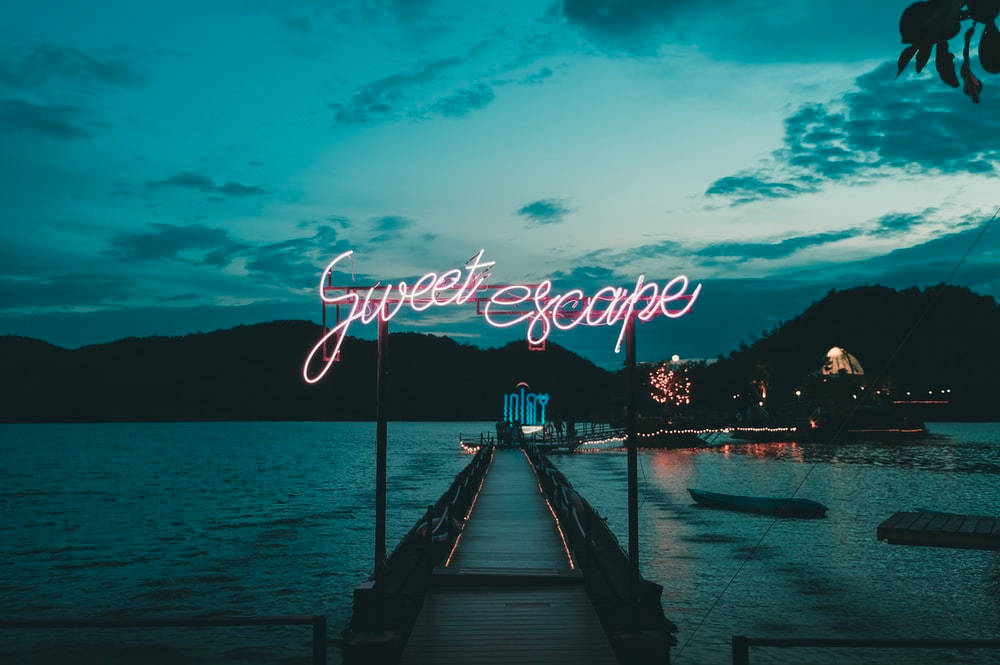 漂亮美學 桌布 壁紙 — 一個發光的粉紅色霓虹燈牌寫著 Sweet Escape，懸掛在延伸至暮色中平靜湖面的木製碼頭上，遠處有山丘和燈光。