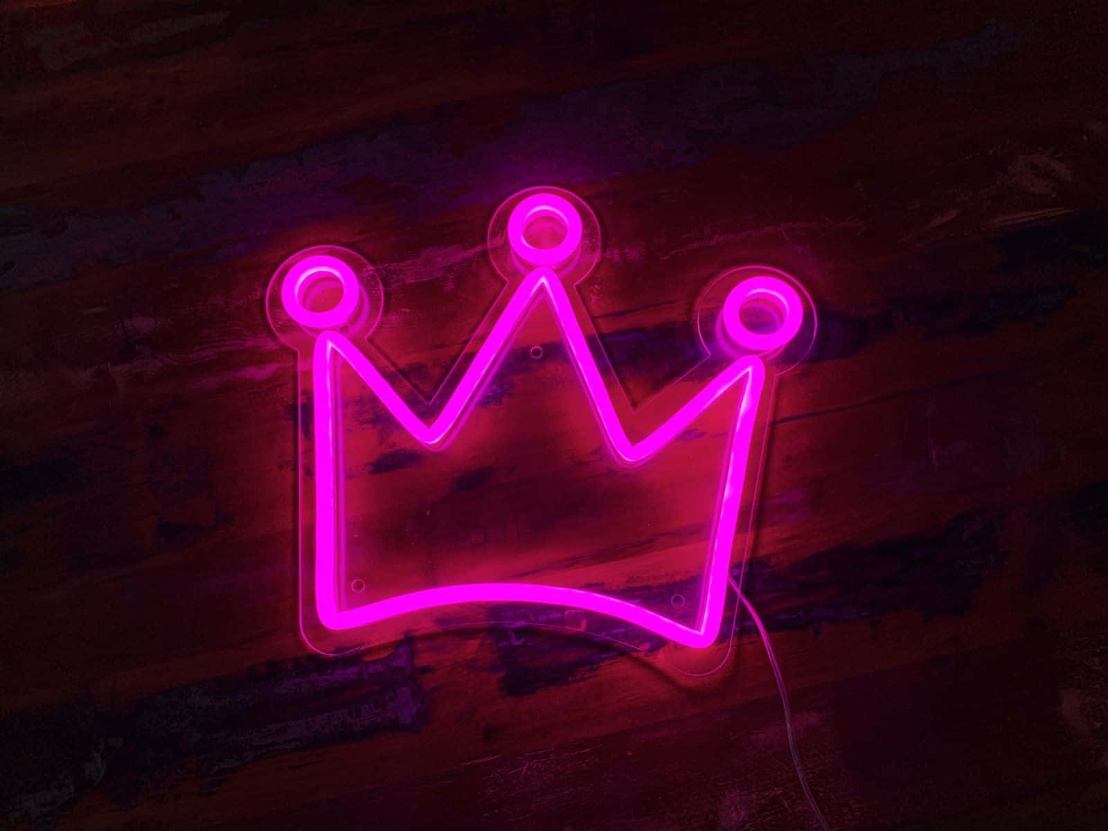 Neon Lights Aesthetic 桌布 壁紙 — 一個發光的粉紅色霓虹皇冠標誌在昏暗的空間中，將鮮豔的光芒投射在深色質樸的木牆上。