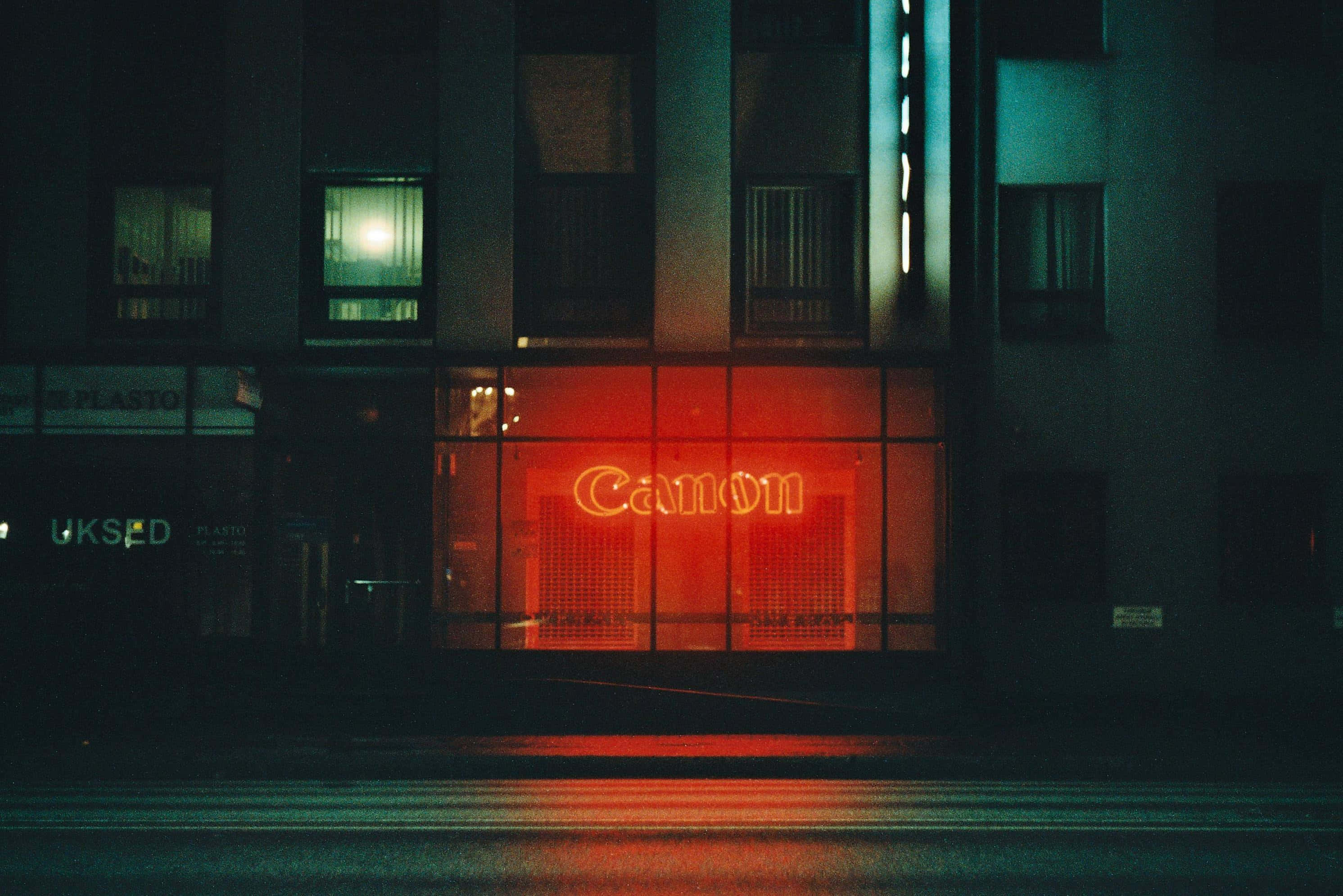 美學 桌布 壁紙 — 一個發光的紅色Canon霓虹燈牌在夜間照亮濕漉漉的城市街道，在黑暗建築物外牆下的人行道上投射出倒影。