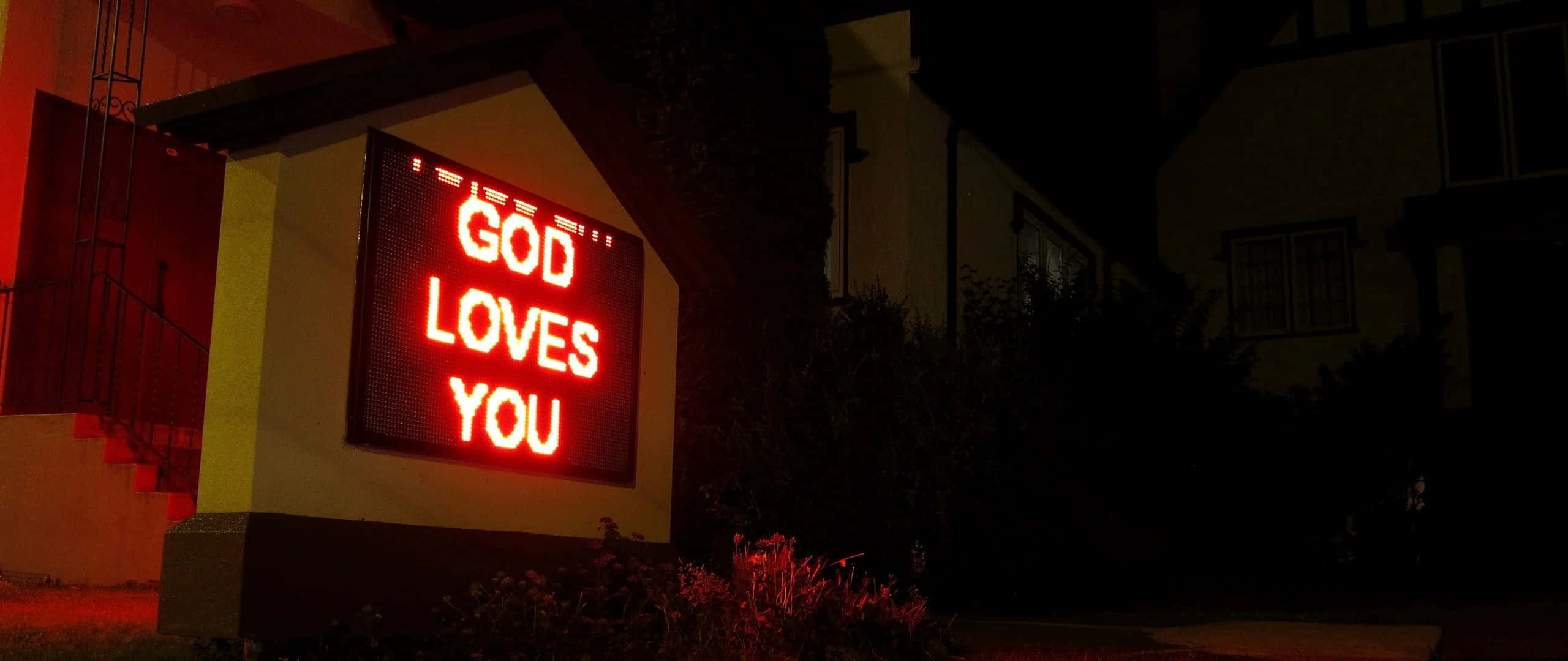 God Loves You 桌布 壁紙 — 一個發光的紅色LED標誌，寫著「神愛你」，在夜間照亮了一棟黑暗的郊區房屋。