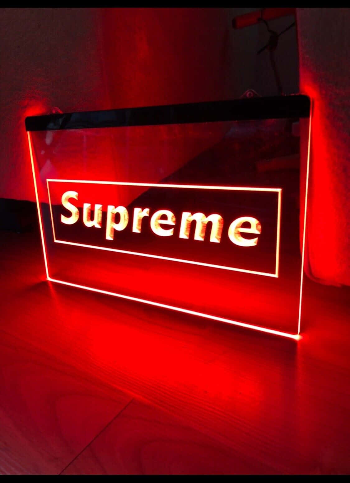supreme 桌布 壁紙 — 一個發光的紅色Supreme標誌招牌在昏暗的房間中將鮮豔的光線投射到木質表面上。