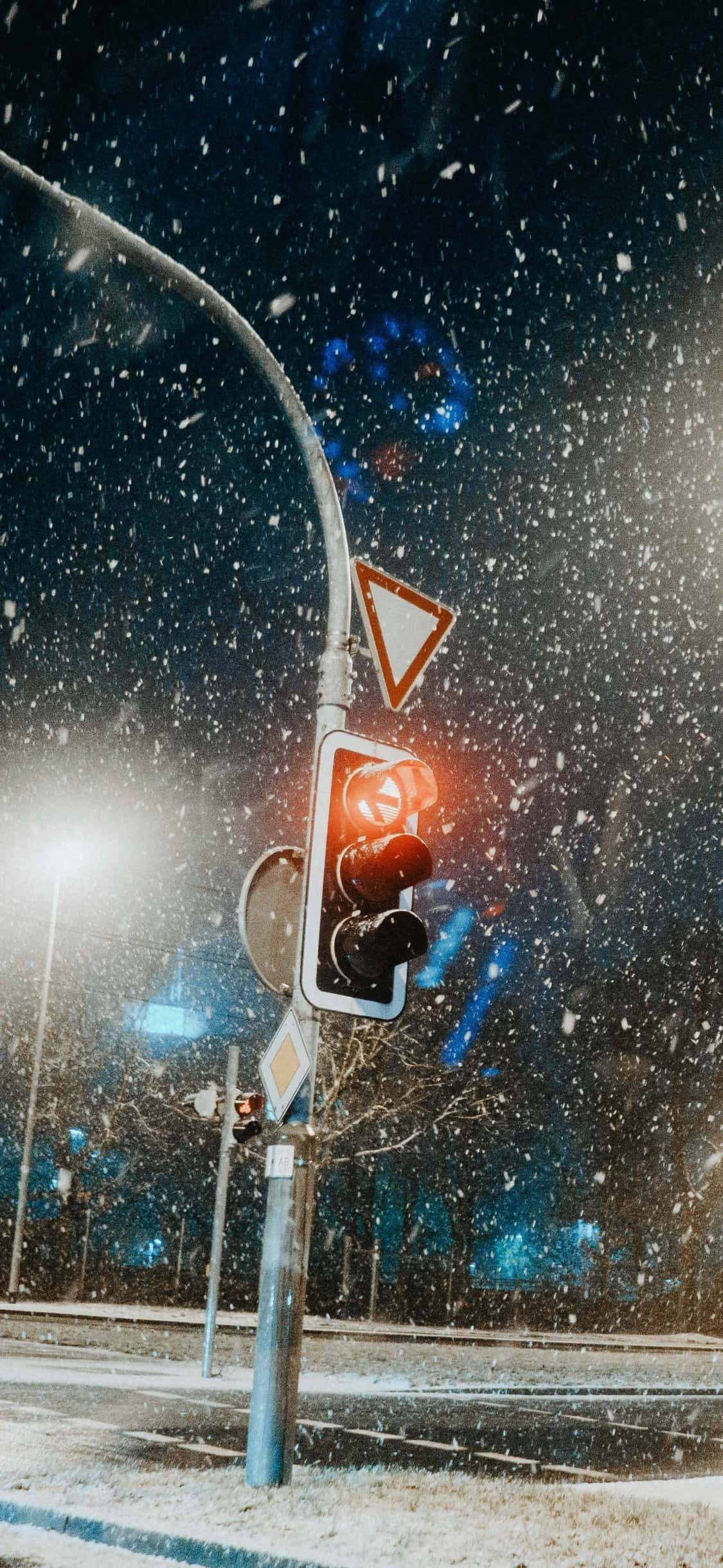 雪 桌布 壁紙 — 一個發光的紅色交通燈獨自矗立在雪景城市夜景中，背景有飄落的雪花和模糊的城市燈光。
