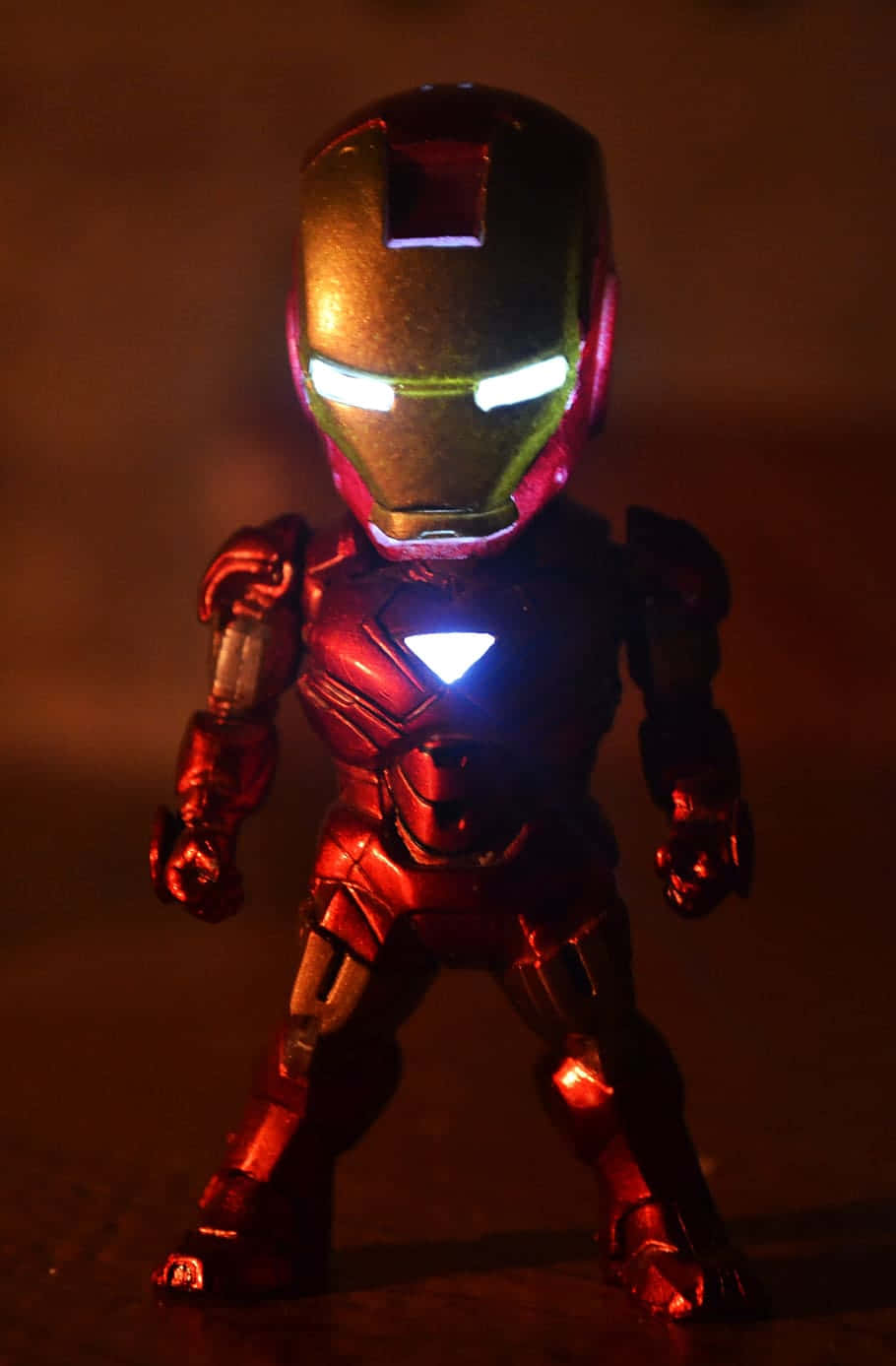 Iron Man Bobbleheads 桌布 壁紙 — 一個發光的紅色和金色鋼鐵人動作人偶站在昏暗的燈光下，眼睛和胸部弧形反應爐發光。