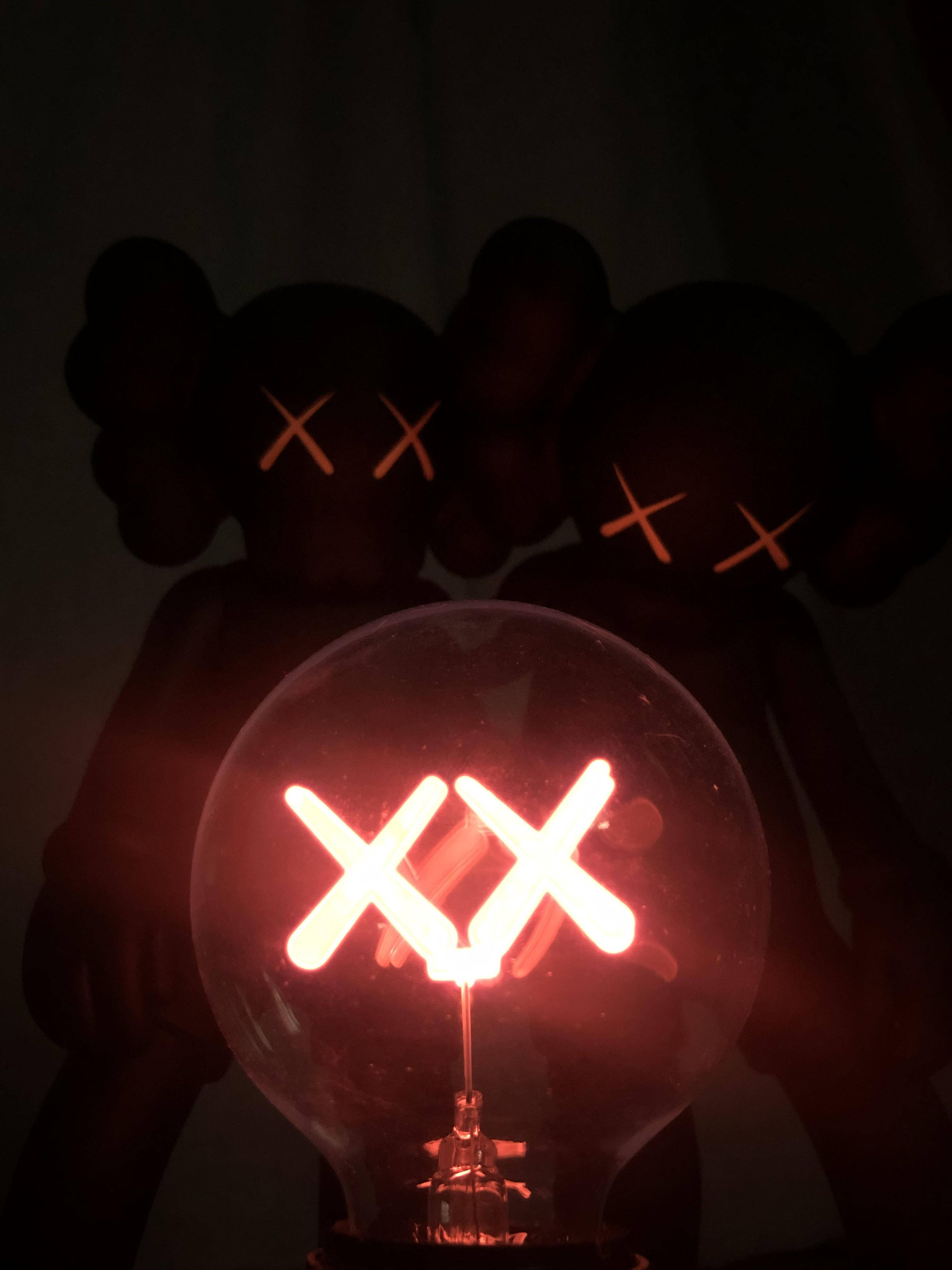 kaws 4K 桌布 壁紙 — 一個發光的紅色燈泡，帶有X形燈絲，在黑暗中將溫暖的光芒投射到其後方的KAWS剪影人物上。