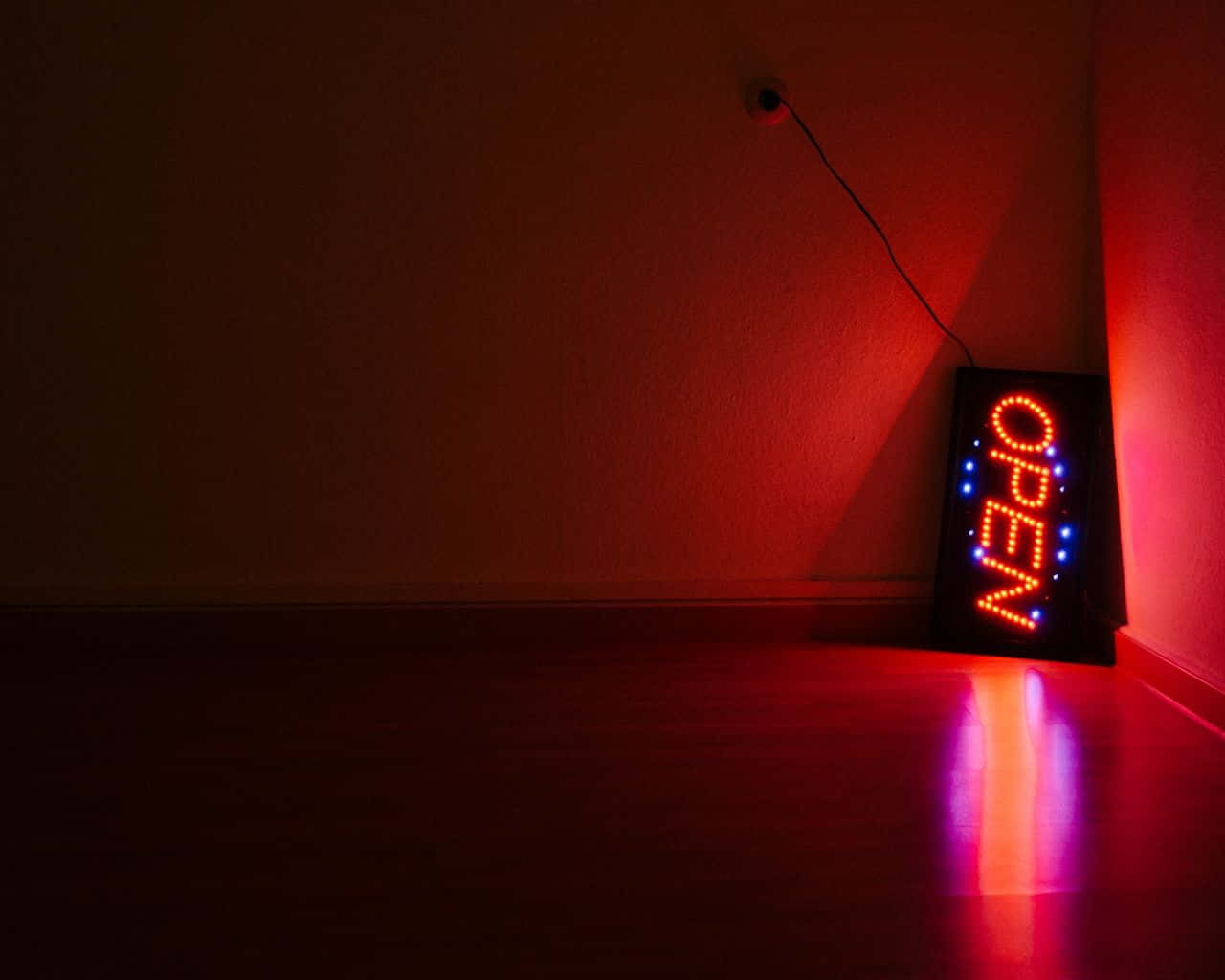 Red Neon 桌布 壁紙 — 一個發光的紅色營業中標誌在昏暗的角落將溫暖的光線投射在深色木地板和紋理牆壁上。