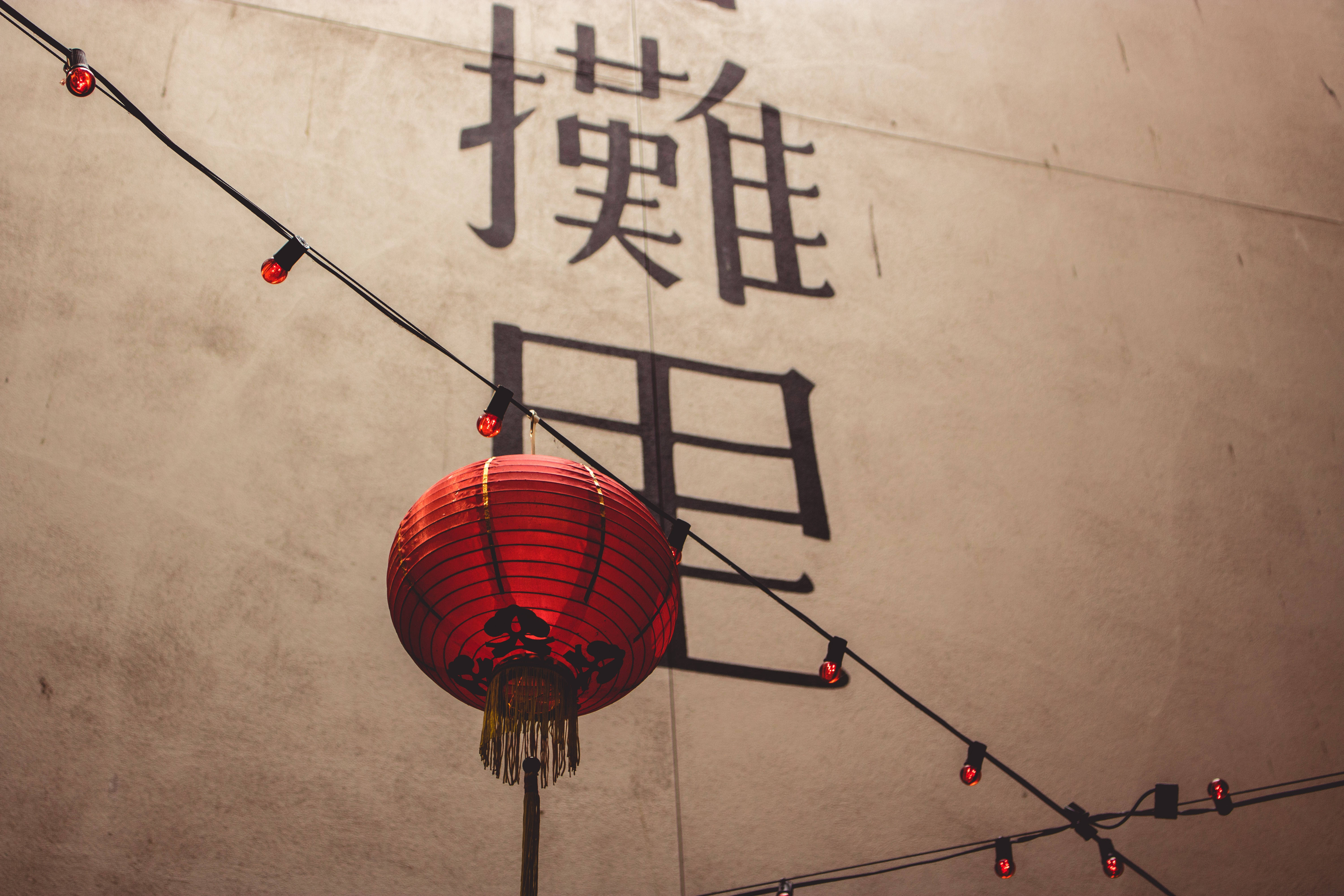 中文 桌布 壁紙 — 一個發光的紅色紙燈籠掛在米色牆上，牆上有大型黑色中文字和燈串。