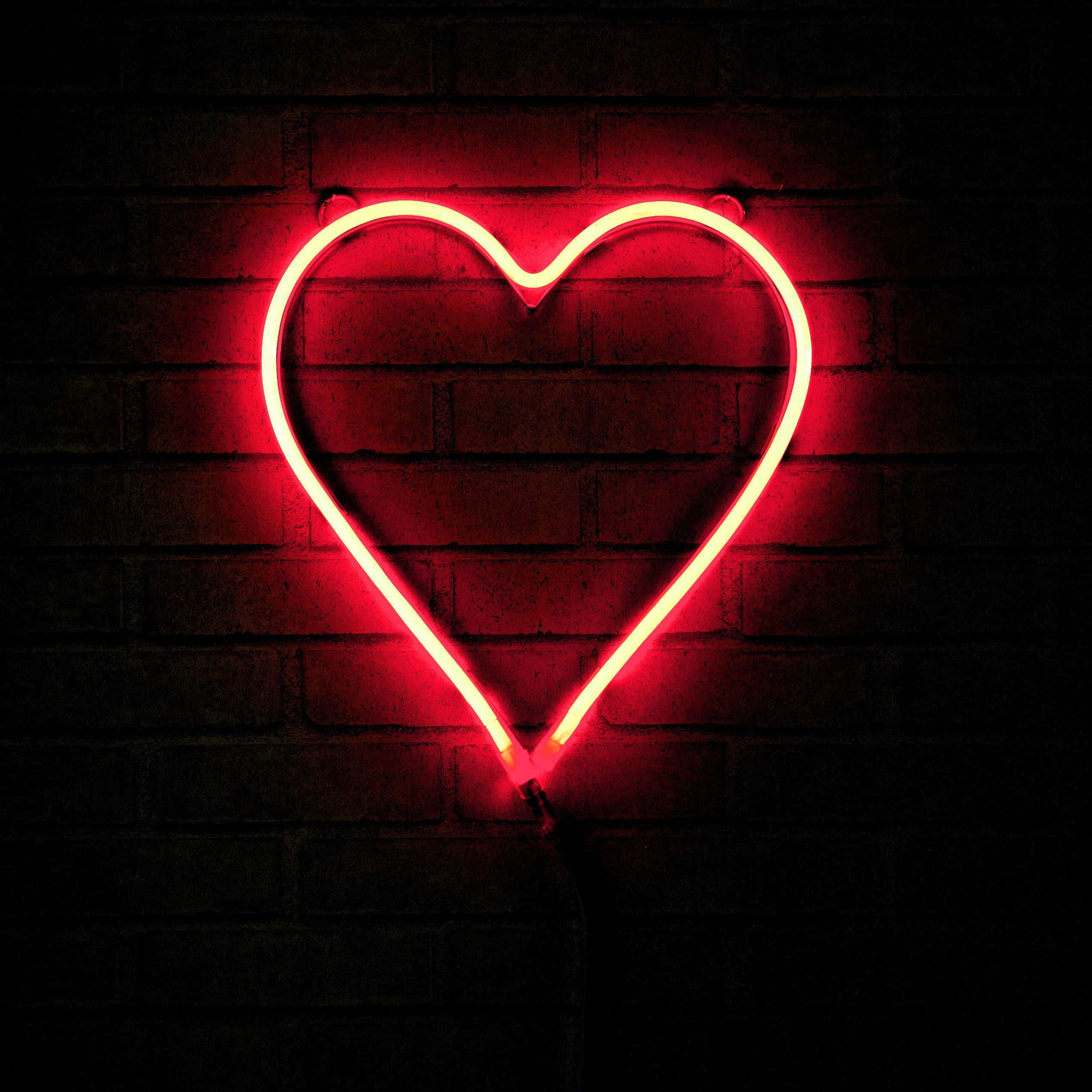 Heart Shape 桌布 壁紙 — 一個發光的紅色霓虹心形在昏暗的空間中，將溫暖的光線投射在深色磚牆上。