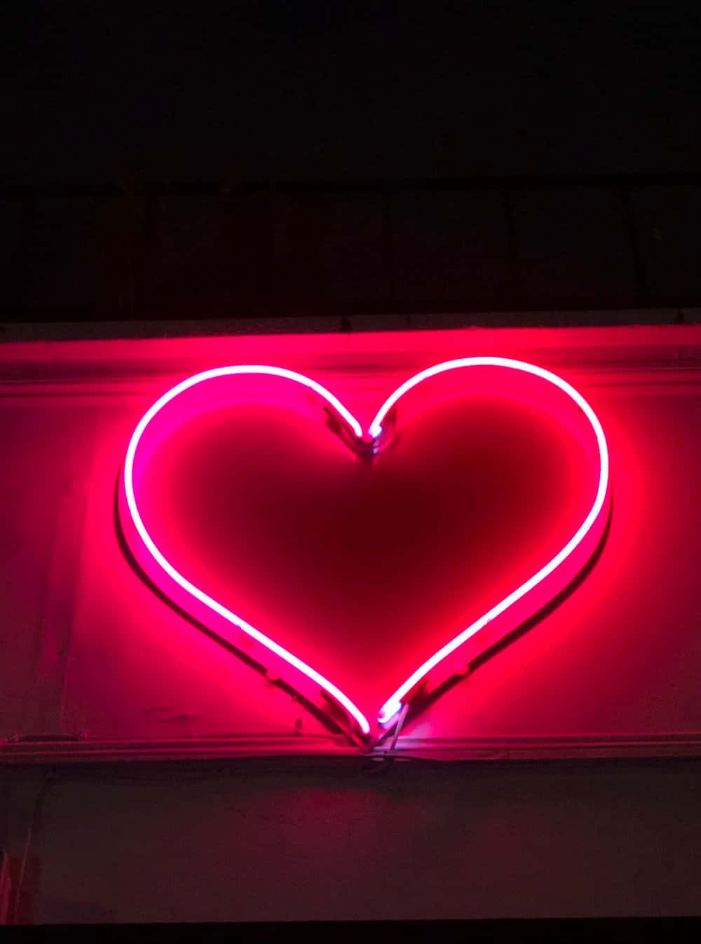 Neon Light 桌布 壁紙 — 一個發光的紅色霓虹心形標誌在夜間城市環境中，在黑暗的牆壁上投射出溫暖的光芒。