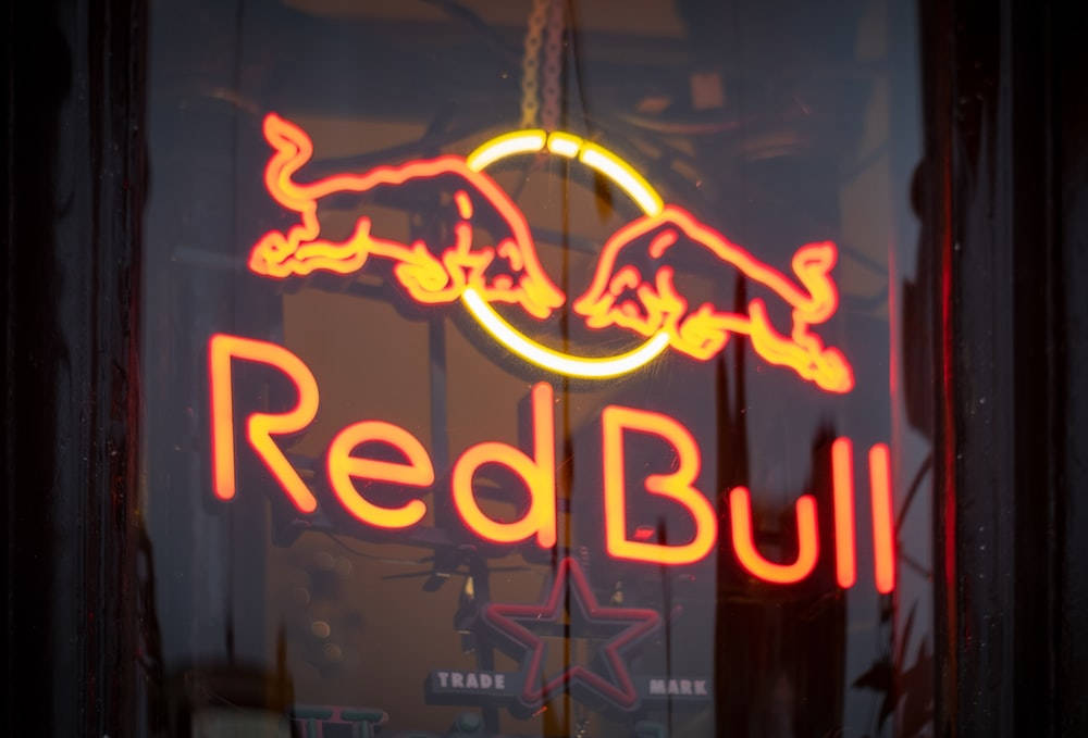 Red Bull 桌布 壁紙 — 一個發光的紅色霓虹燈標誌，兩頭衝鋒的公牛環繞著Red Bull字樣，位於昏暗的窗戶中。
