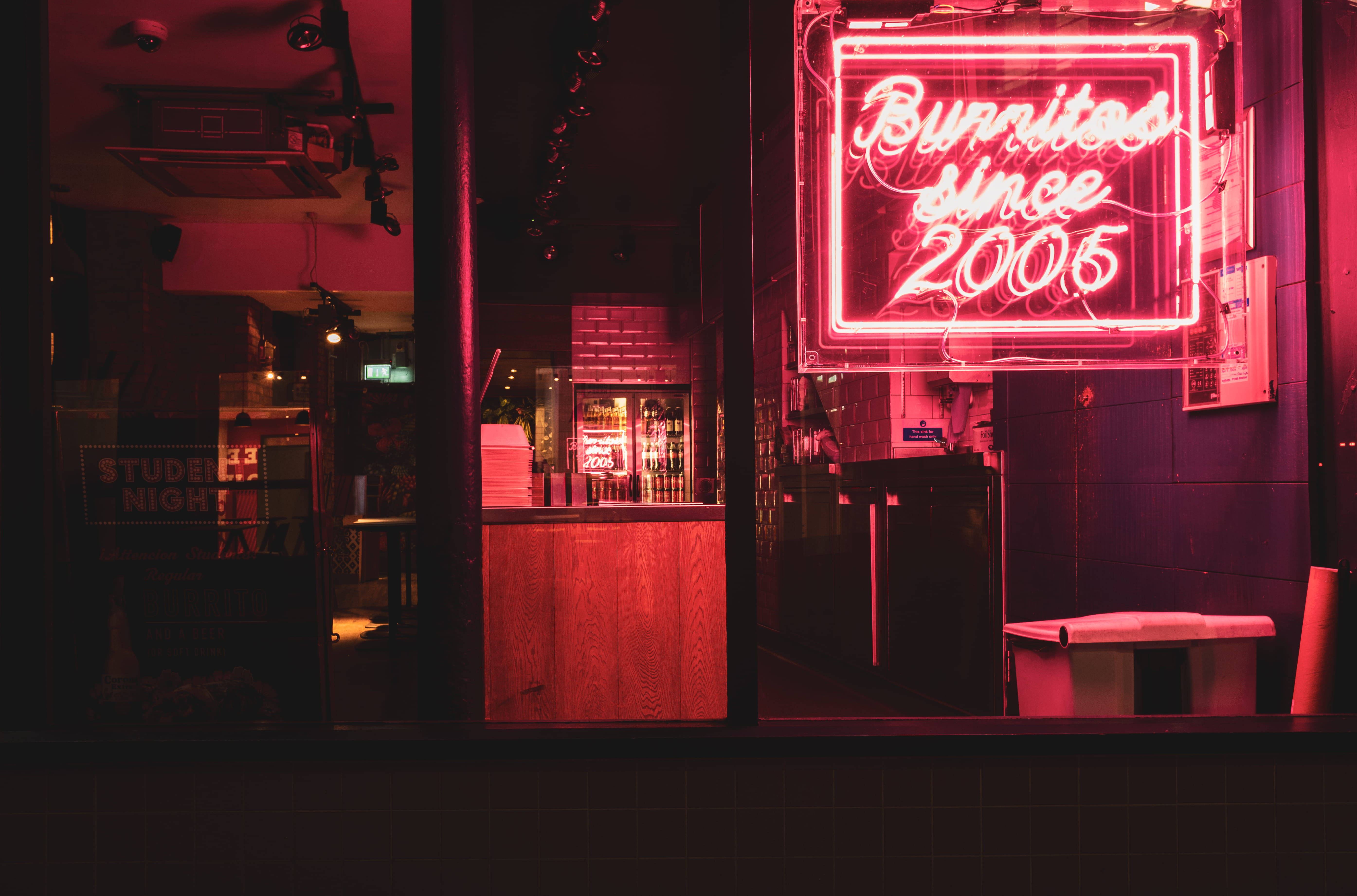 墨西哥捲餅 桌布 壁紙 — 一個發光的紅色霓虹燈牌，上面寫著「Bunnies since 2005」，在夜間透過窗戶照亮昏暗的酒吧。