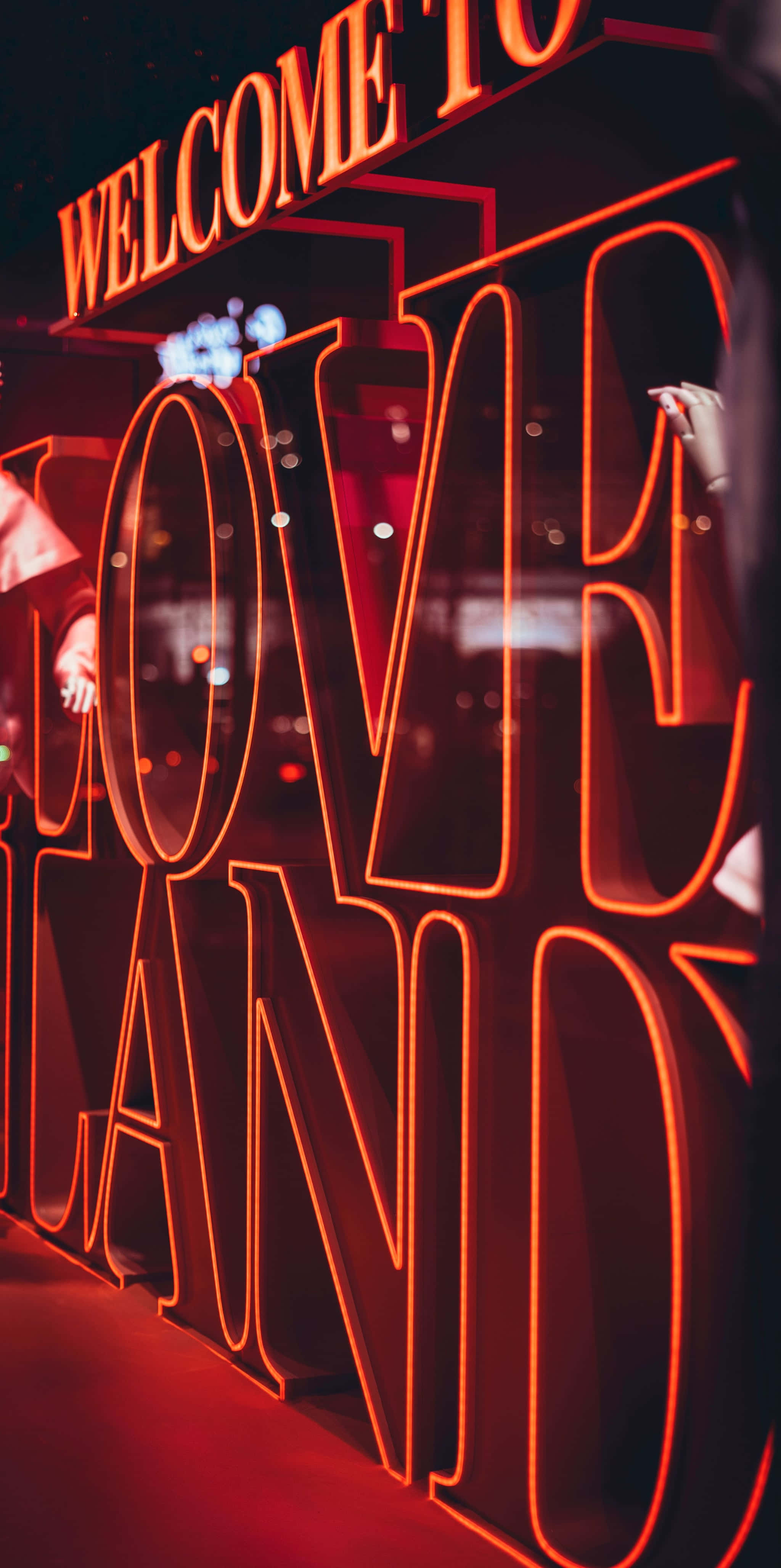Welcome Sign 桌布 壁紙 — 一個發光的紅色霓虹燈牌，上面寫著「WELCOME TO LOVE LAND」，在反射表面上投射出溫暖的光芒，背景有模糊的人影。