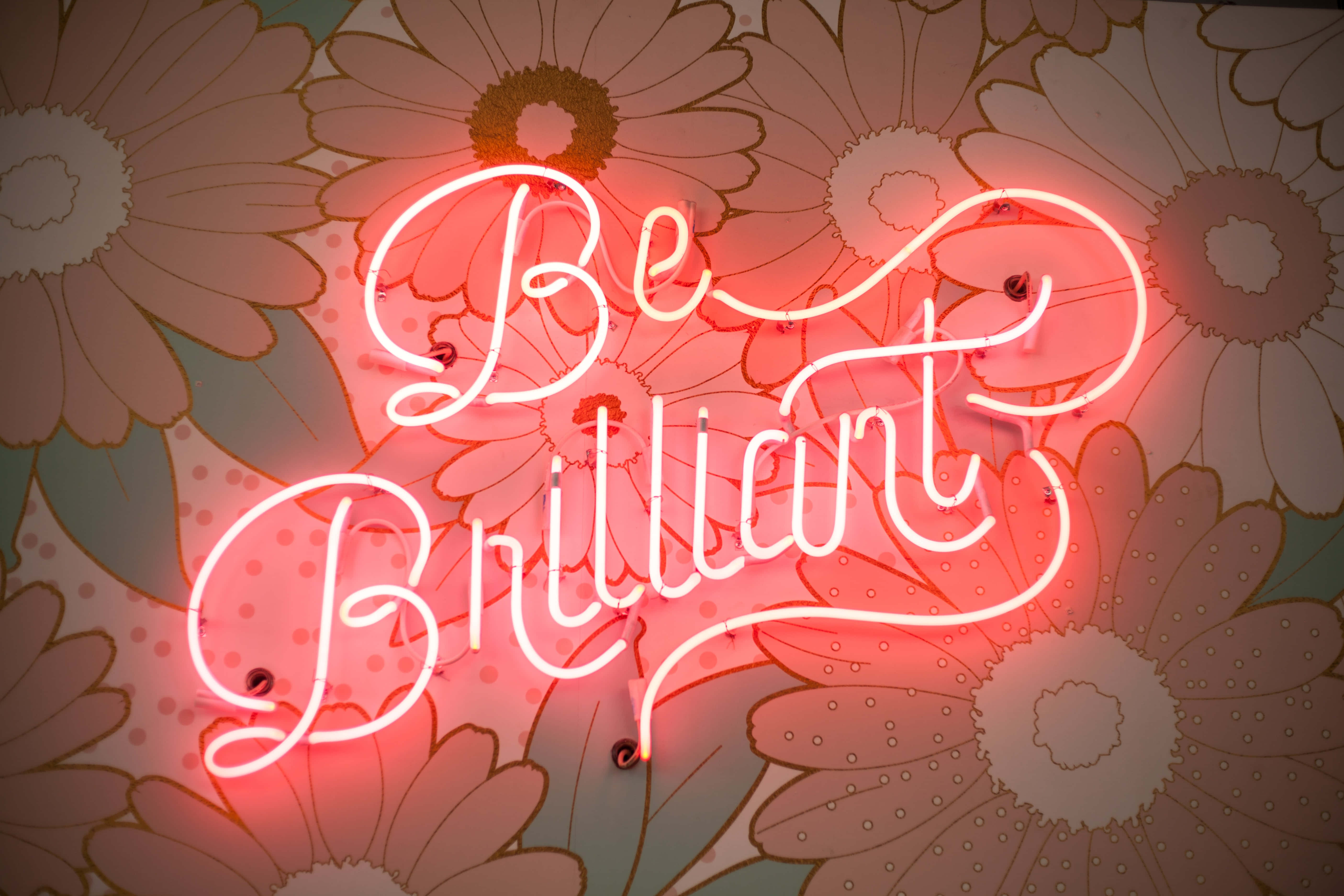 桌布 壁紙 — 一個發光的紅色霓虹燈牌，上面寫著「Be Brilliant」，在柔和粉彩色的花卉壁紙背景下閃耀。